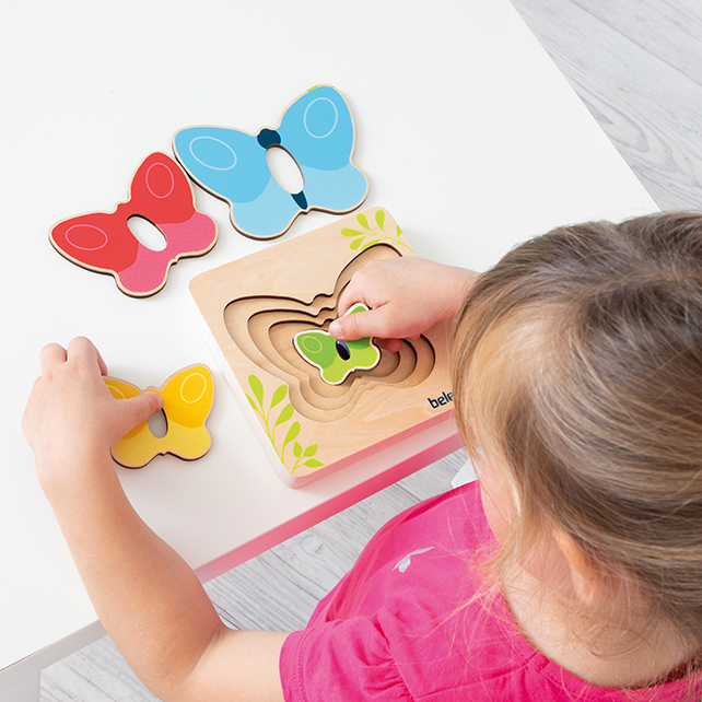 Un niño juega en una mesa con un rompecabezas de mariposa de madera colocándolas piezas de colores.