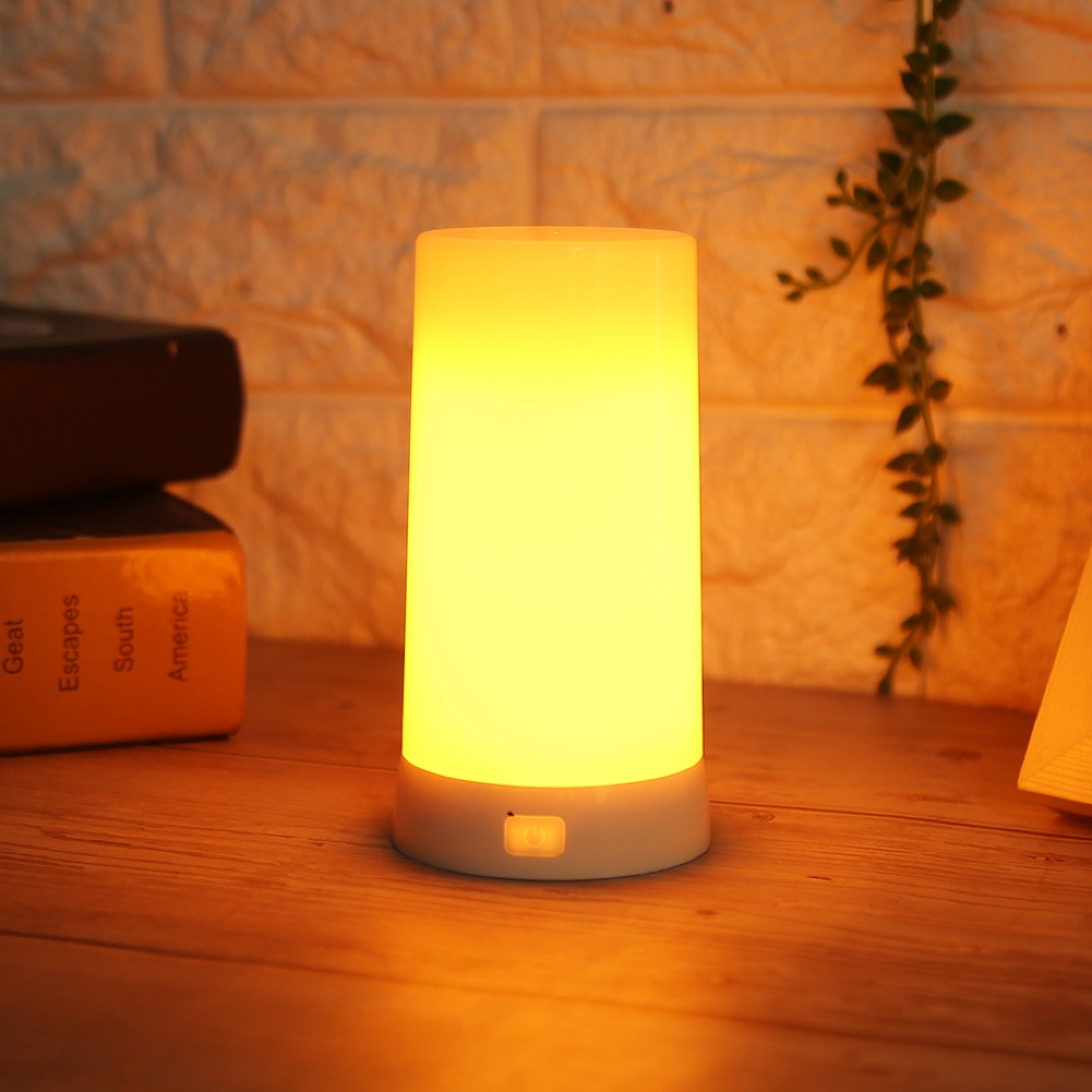 Eine leuchtende zylindrische Nachttischlampe auf einem Holztisch mit Büchern und einer Pflanze im Hintergrund.