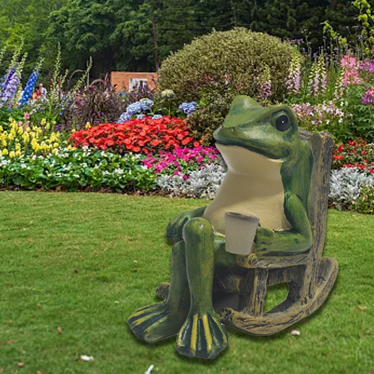 Eine grüne Froschstatue sitzt in einem üppigen Garten mit bunten Blumen auf einem kleinen Schaukelstuhl und hält eine Tasse.