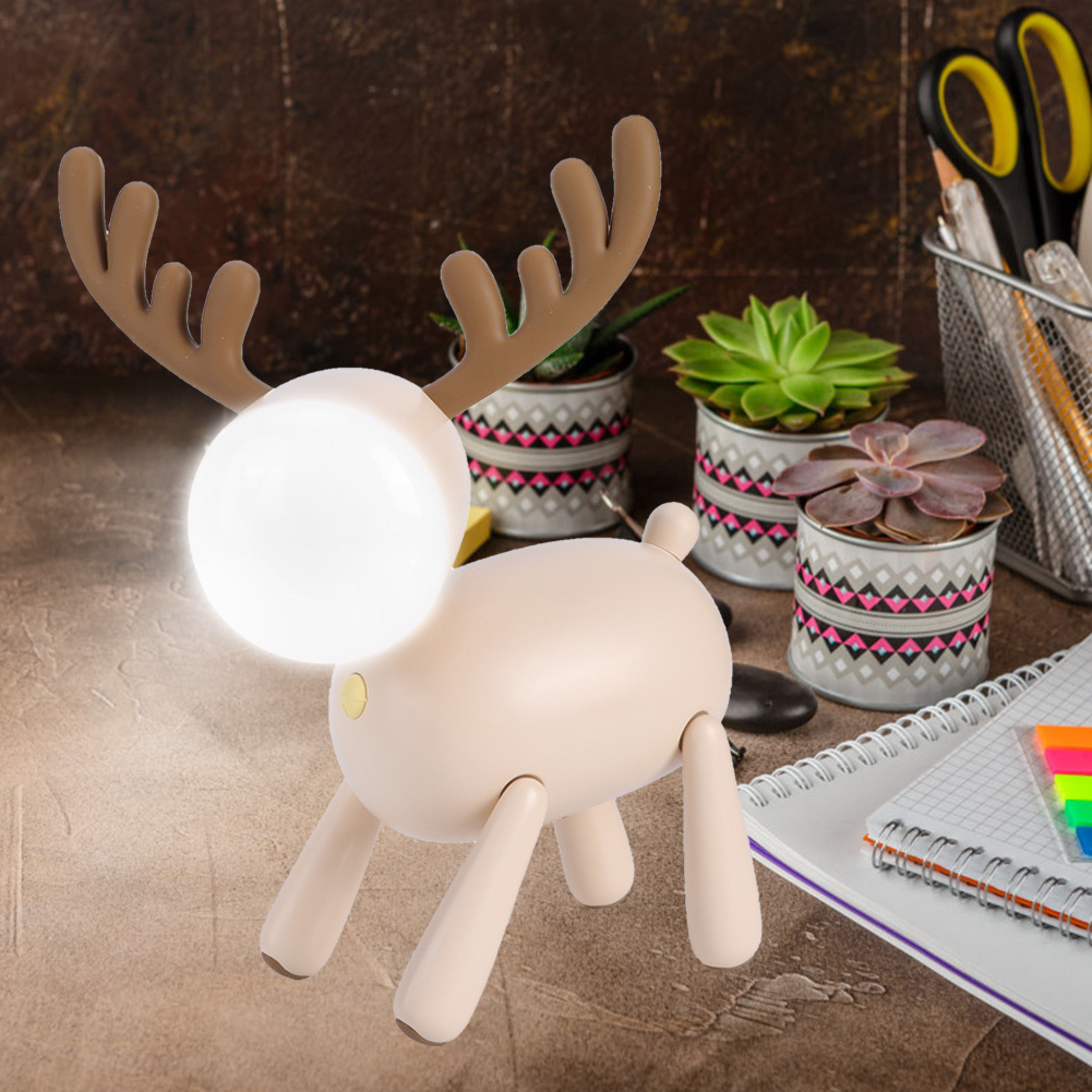 Eine beige Elch-Lampe mit leuchtendem Kopf steht auf einem Tisch neben Notizbüchern und Pflanzen.