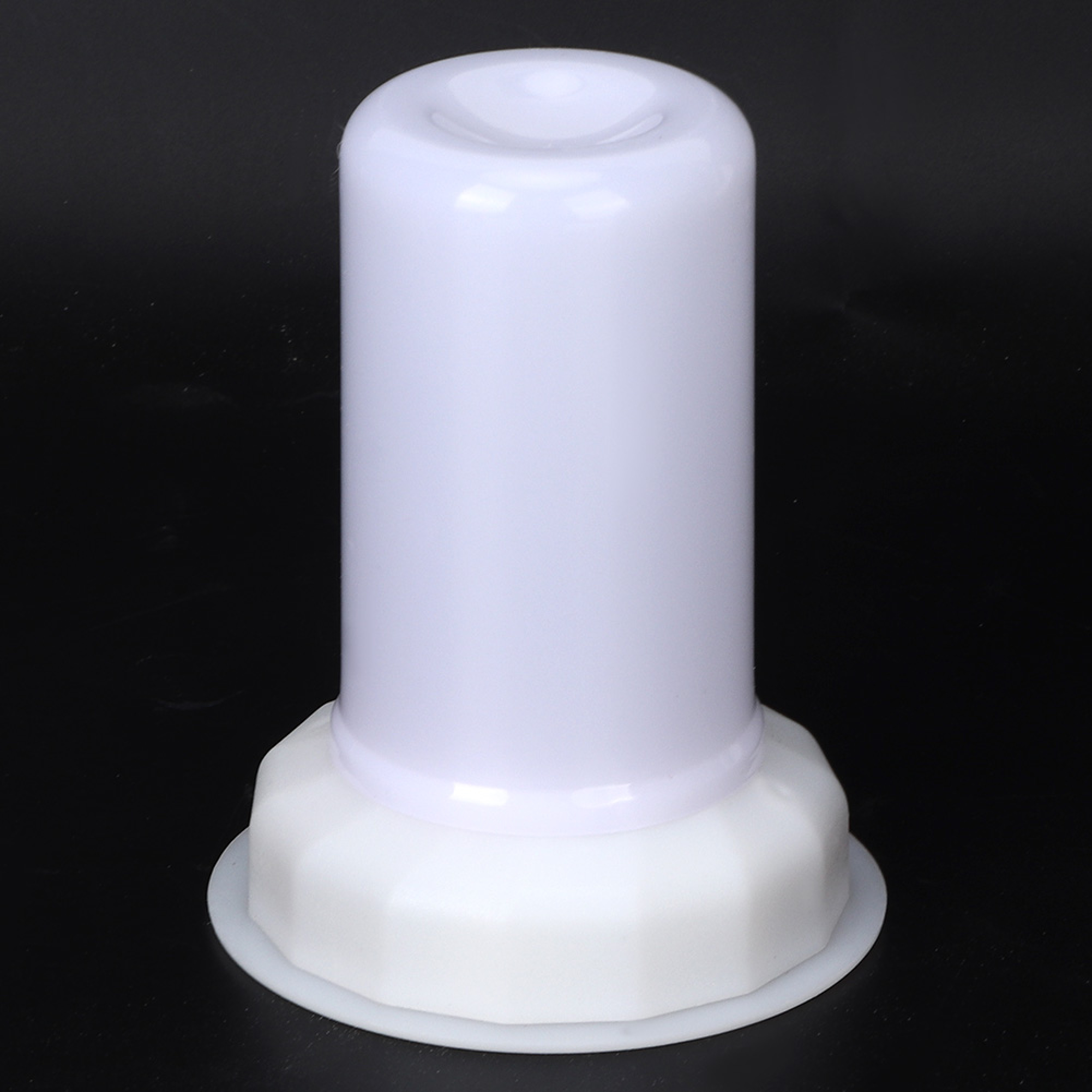 Ein weißes Plastikobjekt mit zylindrischer Oberseite und breiterem, facettierten Sockel vor schwarzem Hintergrund.