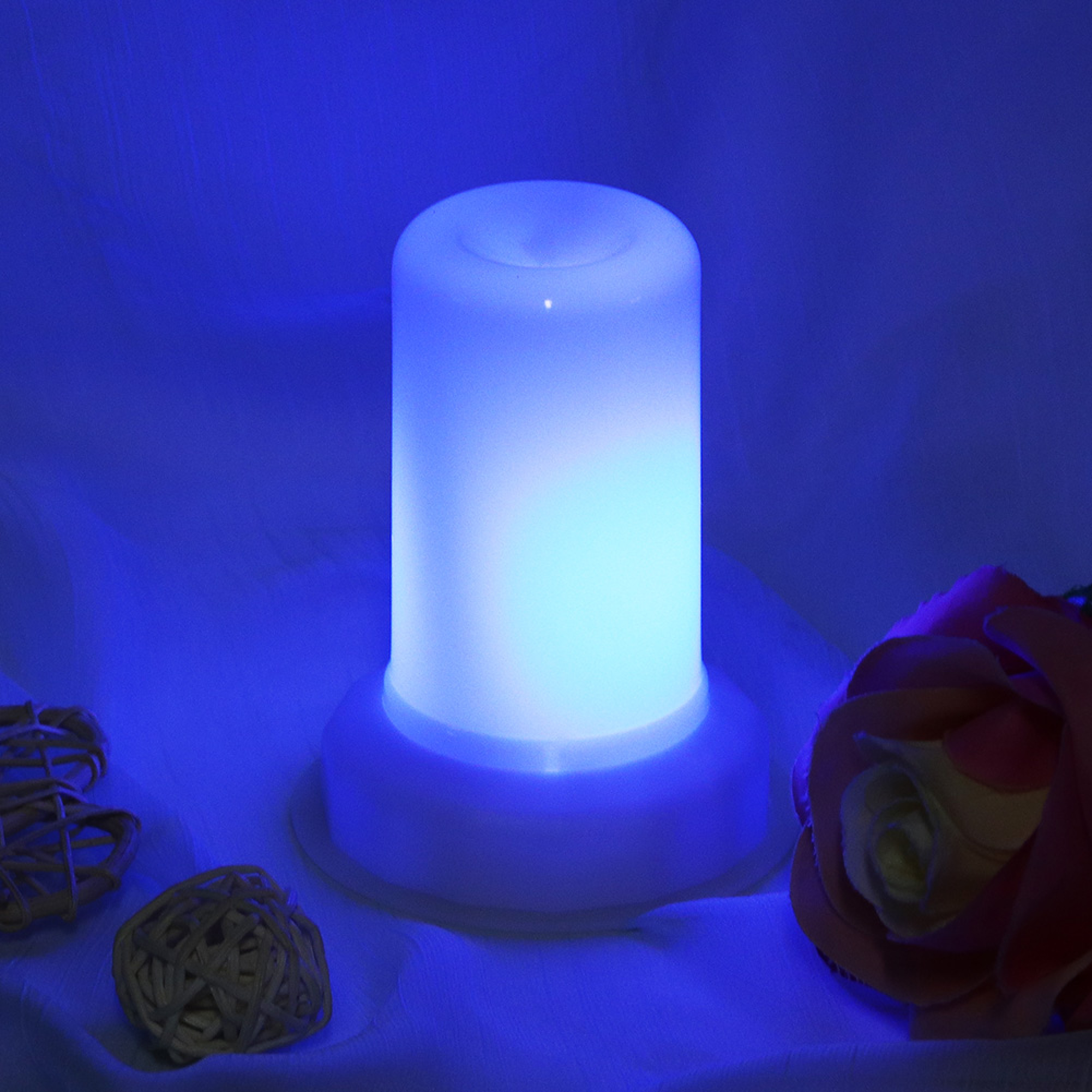 Ein leuchtend blauer Zylinderdiffusor steht auf einer weißen Oberfläche, flankiert von dekorativen Weidenkugeln und einer Rose.