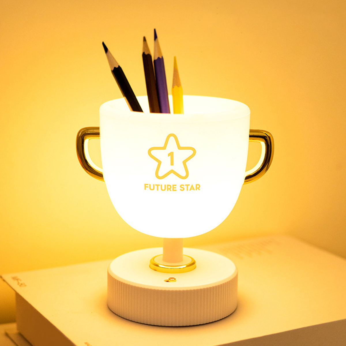 Eine pokalförmige weiße Schreibtischlampe mit Buntstiften steht auf einem weißen Buch vor warmem gelbem Hintergrund.