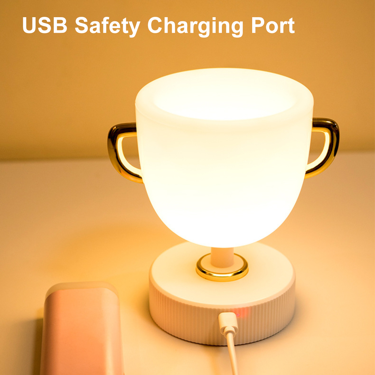 Eine weiß-goldene, tasseförmige Lampe mit USB-Ladeanschluss steht auf einem Tisch vor gelbem Hintergrund.