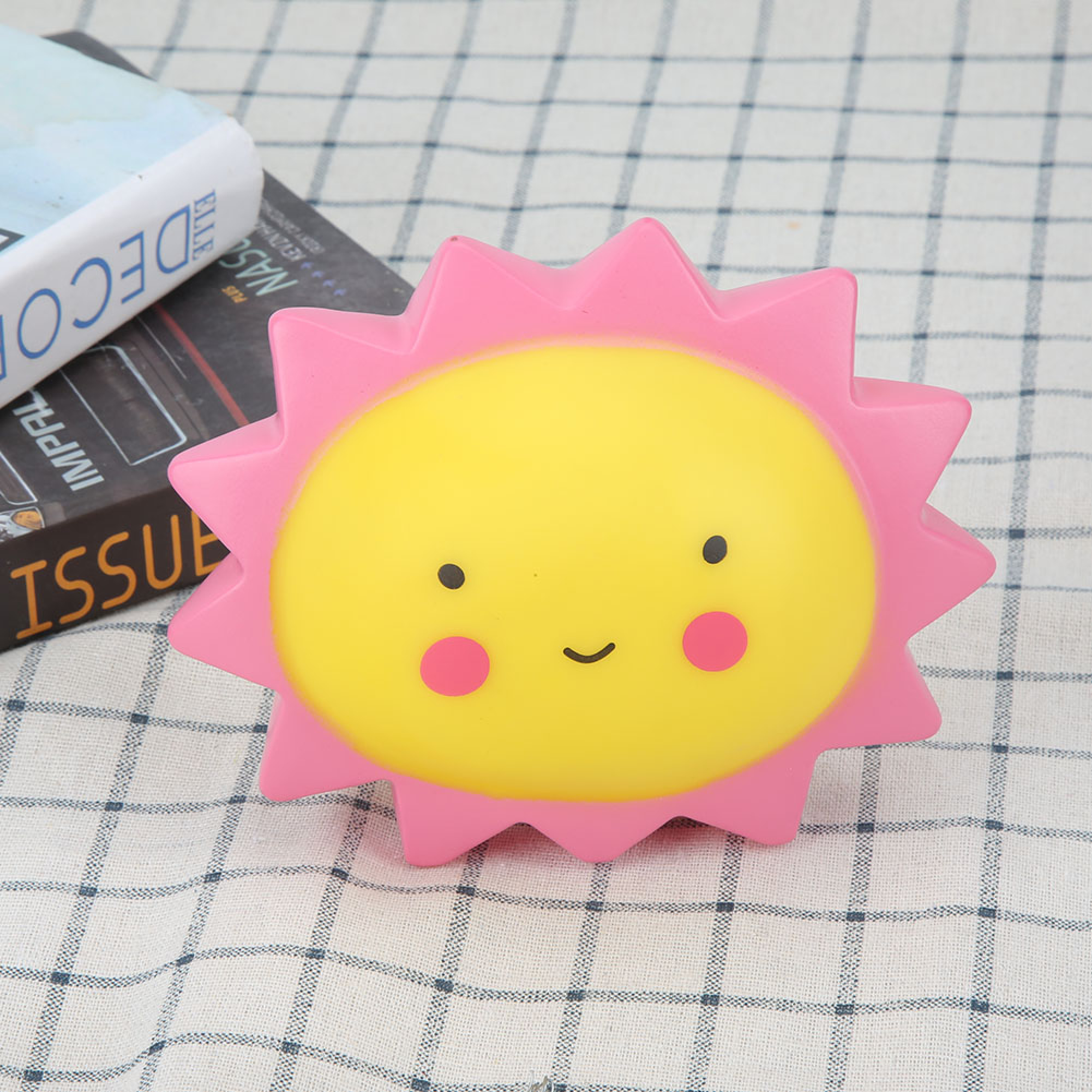 Eine rosafarbene und gelbe Sonnenlampe mit niedlichem Gesicht steht auf einem karierten Tuch neben zwei Büchern.