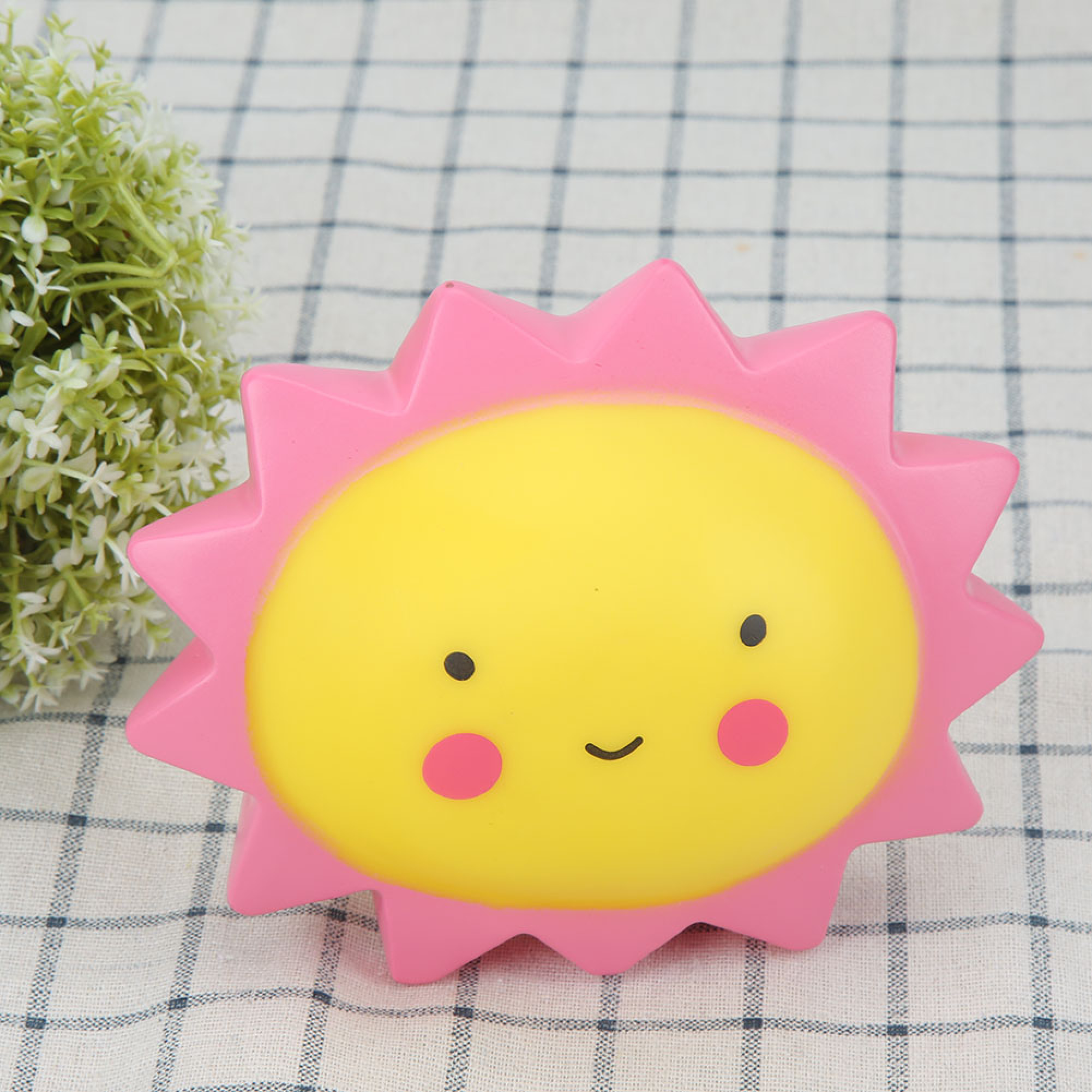 Ein süßes sonnenförmiges Quetschspielzeug auf kariertem Tuch mit einer Pflanze.