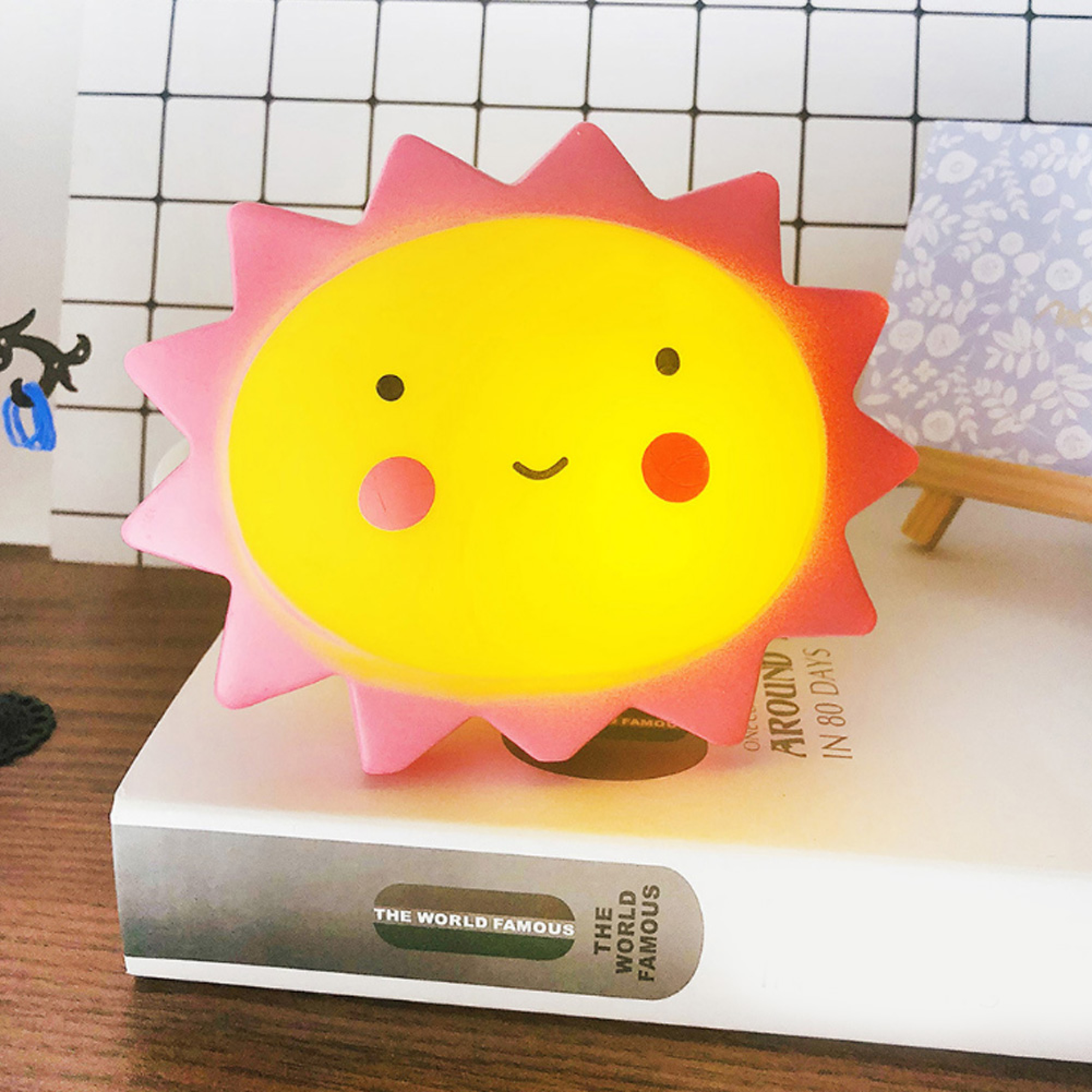 Eine niedliche, gelb-rosa Lampe in Sonnenform auf einem Buch, vor einem karierten Hintergrund.