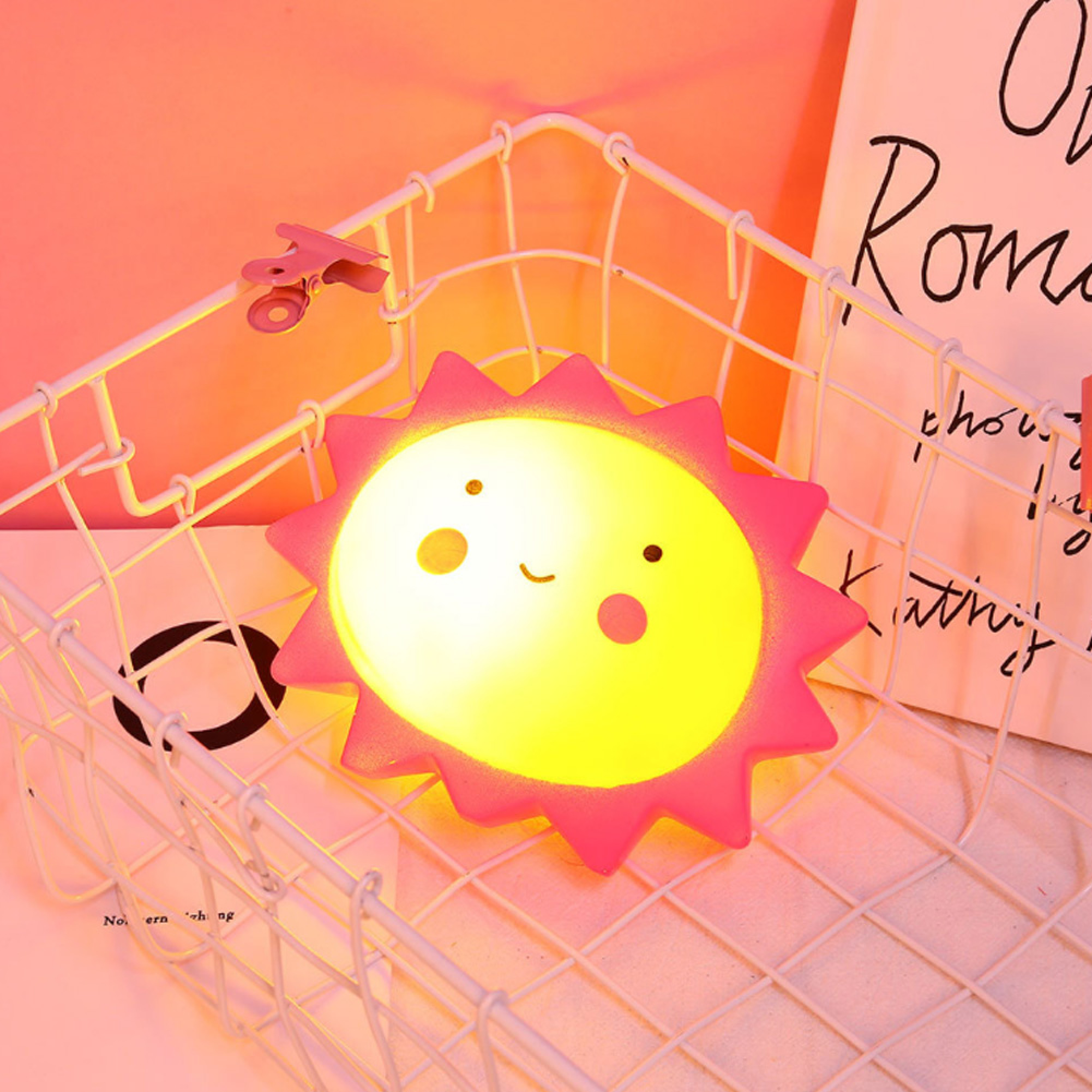 Eine leuchtende, sonnenförmige Lampe mit rosafarbenem Rand liegt in einem weißen Drahtkorb vor einem rosafarbenen Hintergrund.