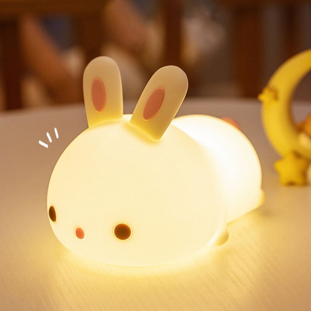 Eine niedliche, leuchtende Hasen-Nachtlampe aus Silikon steht auf einem Tisch, mit einem gelben Mond-Objekt im Hintergrund.