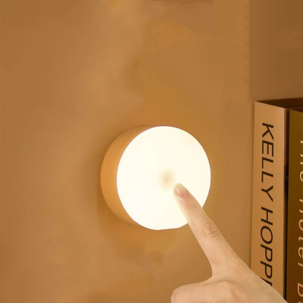Ein Finger berührt eine runde Wandleuchte, die an einer beigefarbenen Wand neben einem Buch leuchtet.