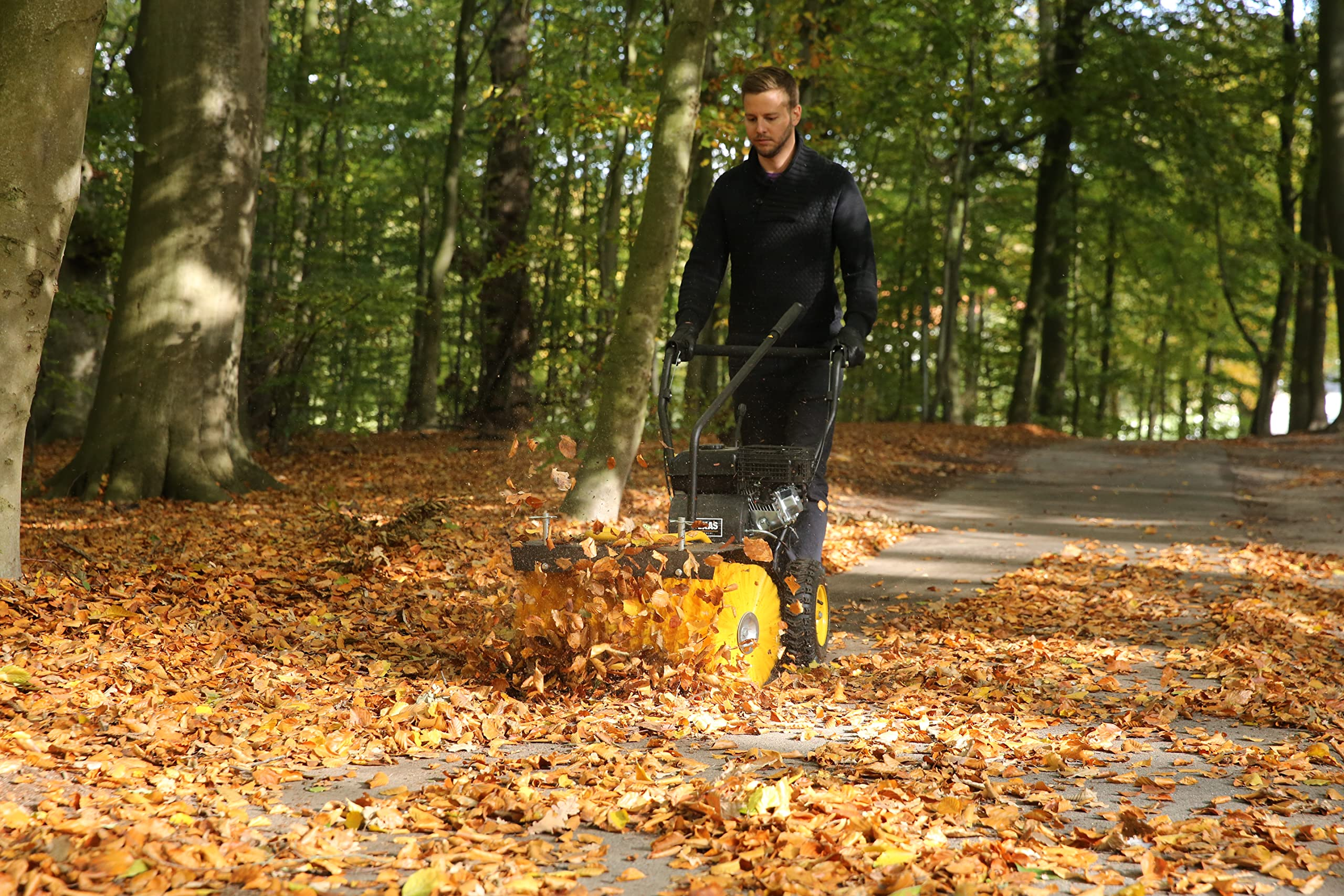 Ein Mann benutzt eine gelbe Kehrmaschine, um trockenes Laub auf einem Waldweg zu räumen.