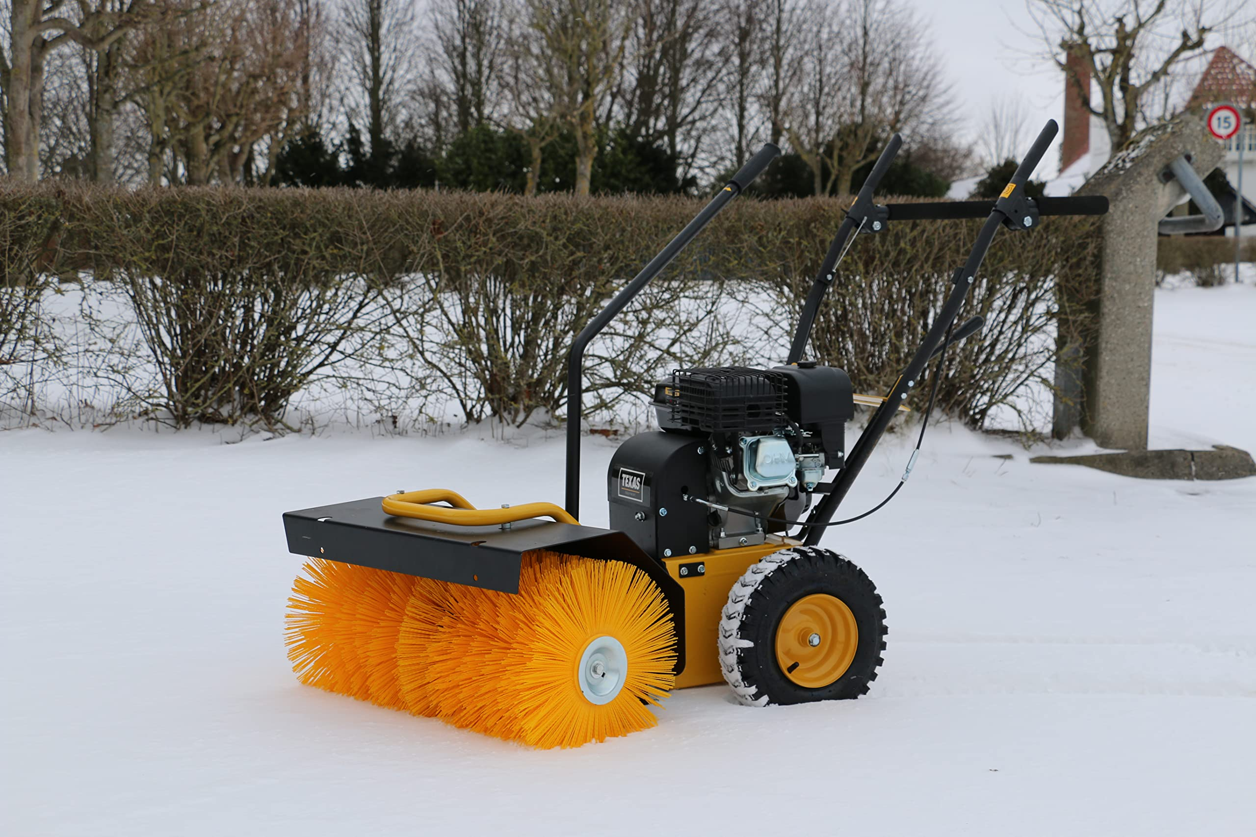 Eine gelbe Motorbürste steht im verschneiten Freien vor einer Hecke.