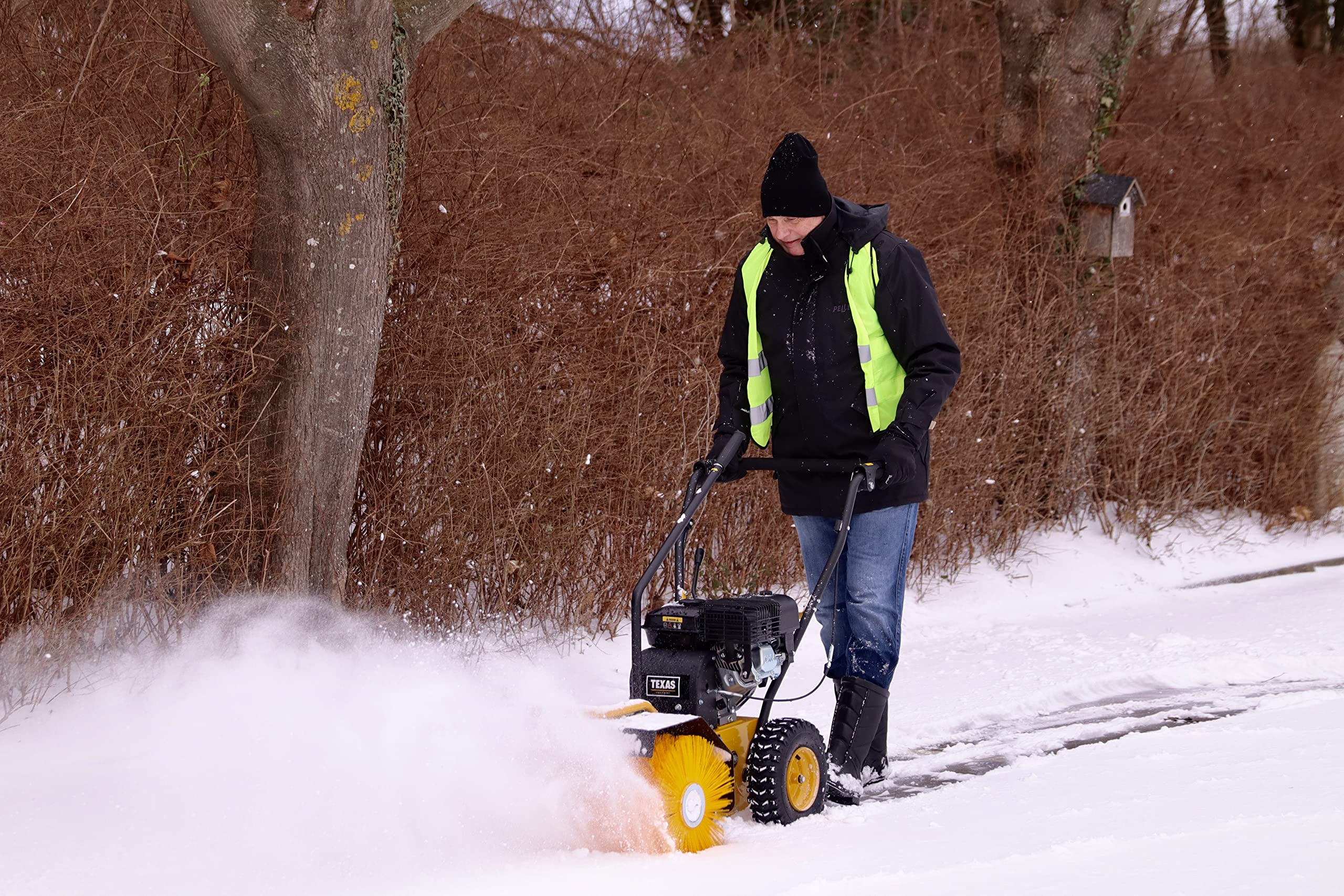 Mann mit schwarzer Jacke und gelber Warnweste kehrt Schnee mit einer gelben Kehrmaschine weg.