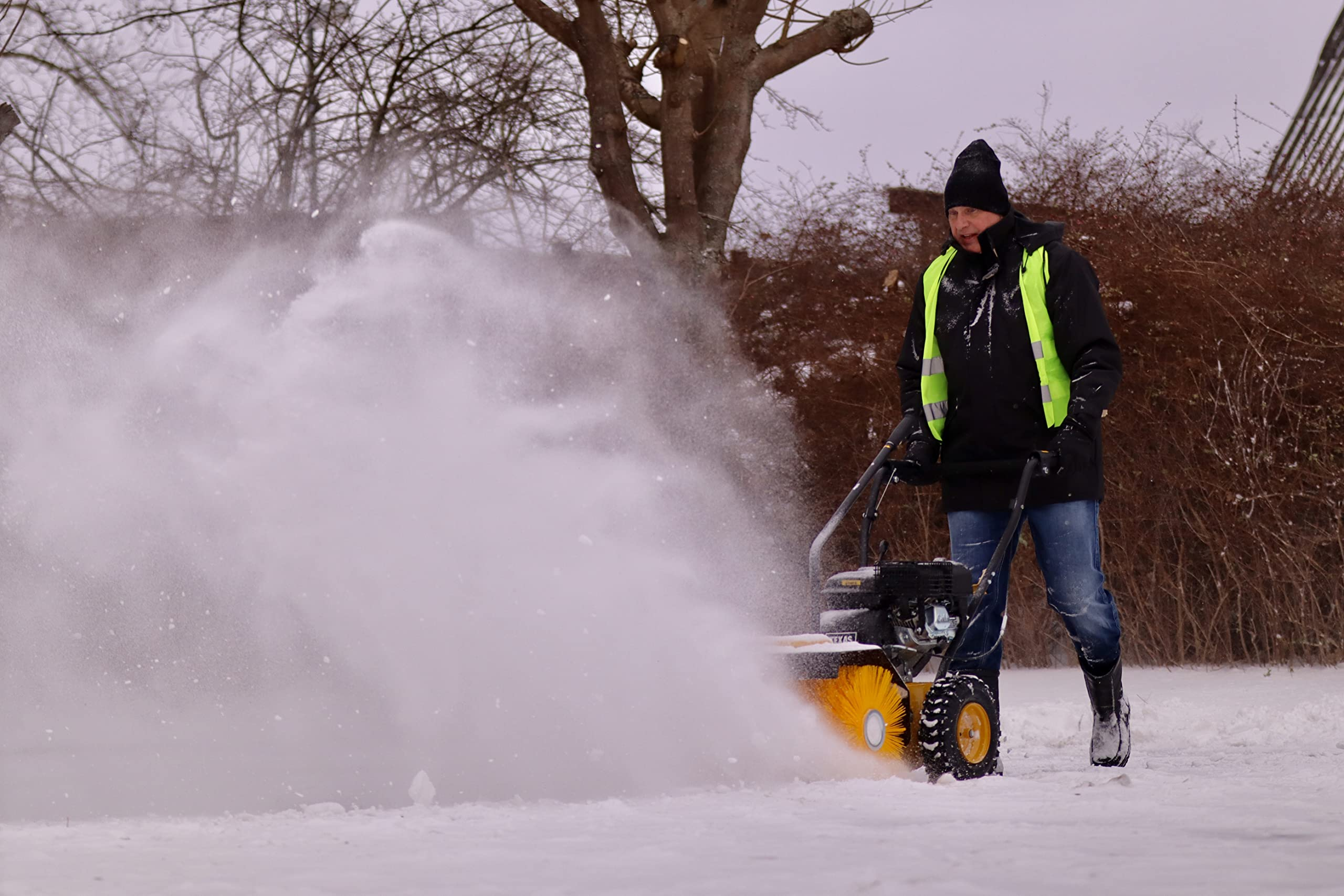 Ein Mann in Jacke und Mütze bedient eine motorisierte Kehrmaschine und wirbelt Schnee auf.
