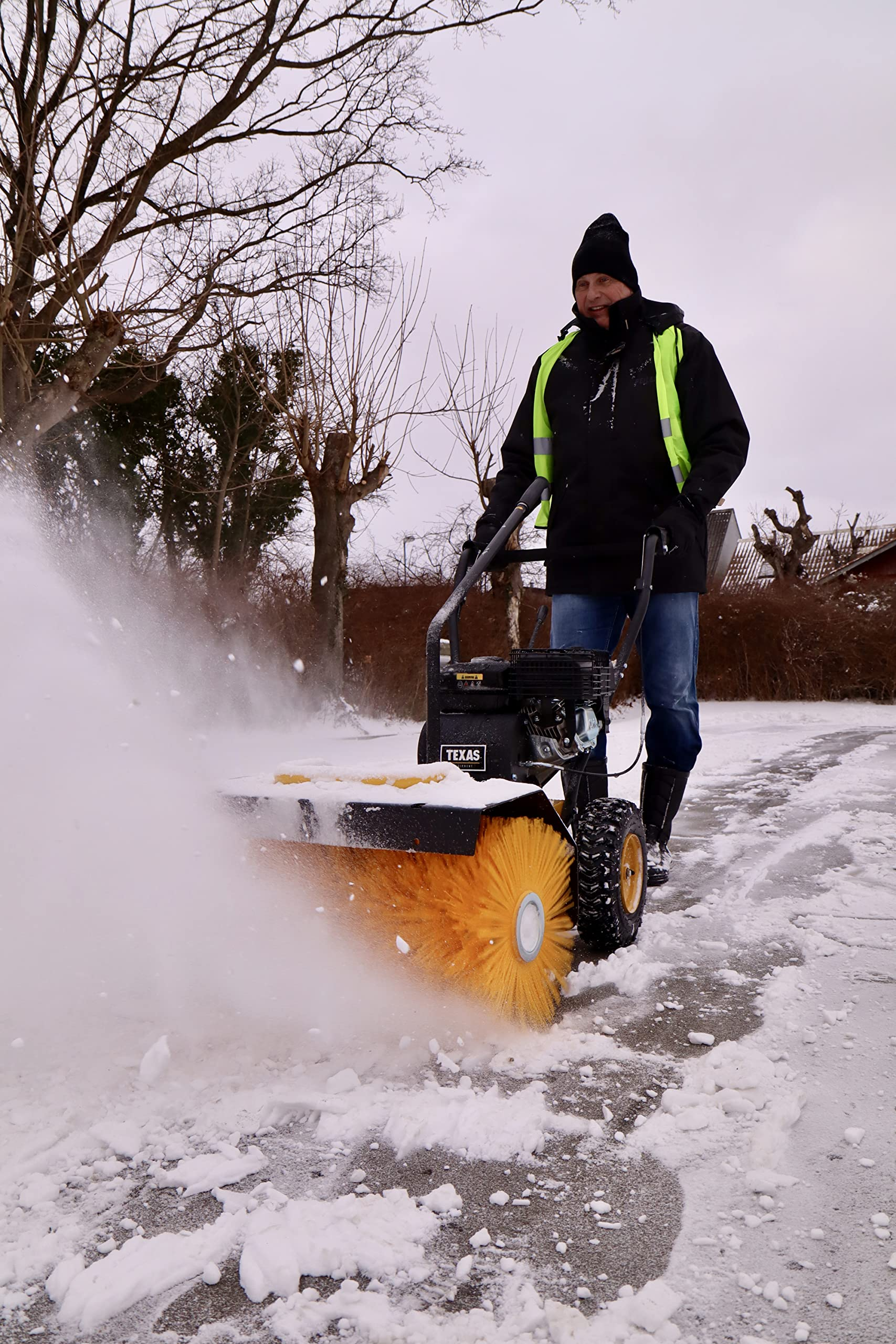 Ein Mann in schwarzer Jacke und Warnweste bedient eine motorisierte Kehrmaschine auf einem verschneiten Weg.