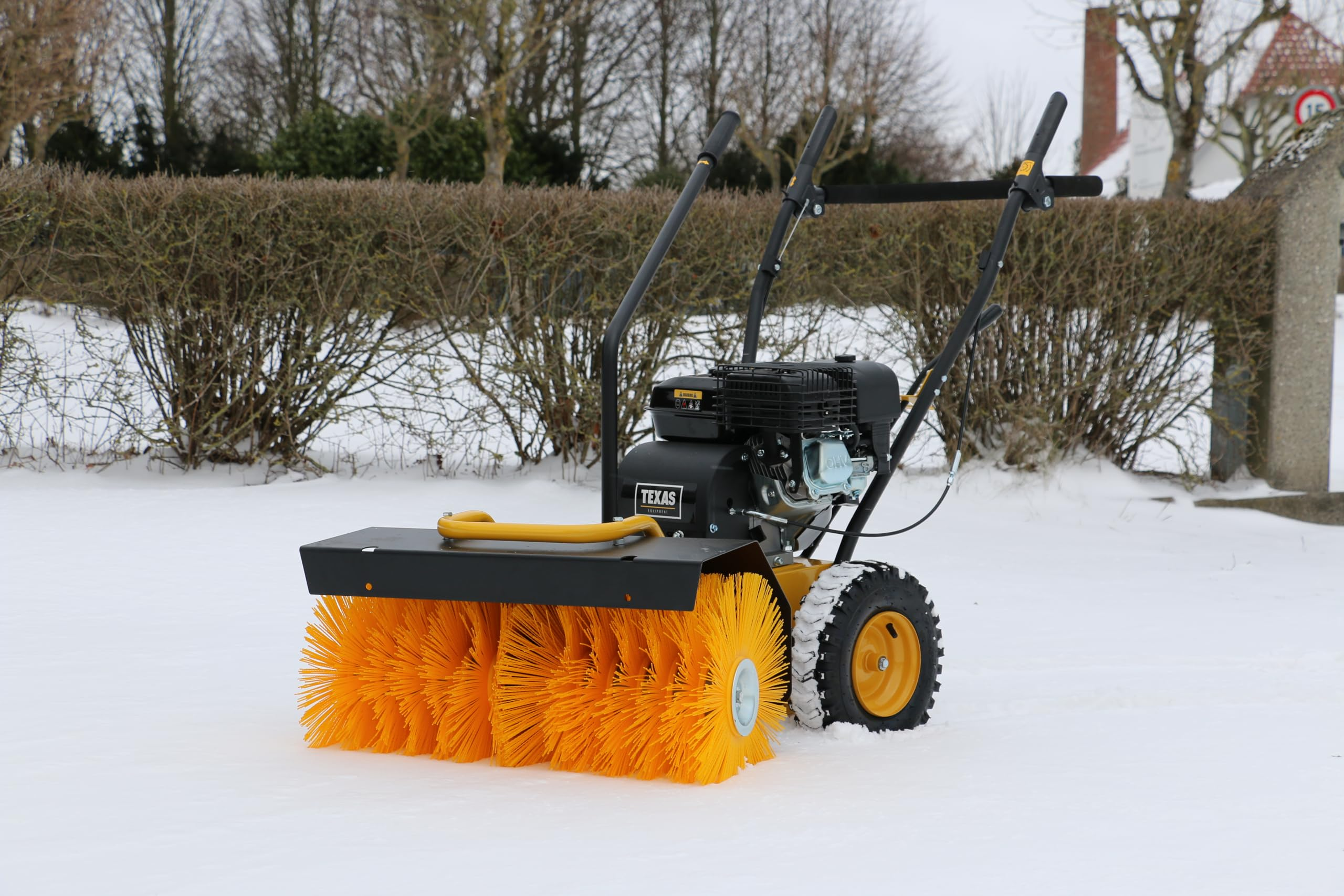 Eine schwarze und gelbe Motorbürste steht im Schnee vor einer Hecke.