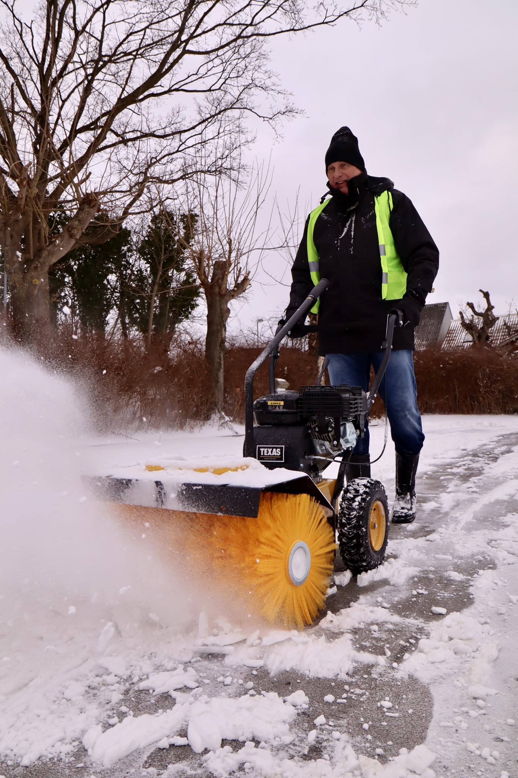 Ein Mann im schwarzen Anzug mit Warnweste bedient eine große gelbe Schneefräse auf einem verschneiten Weg.