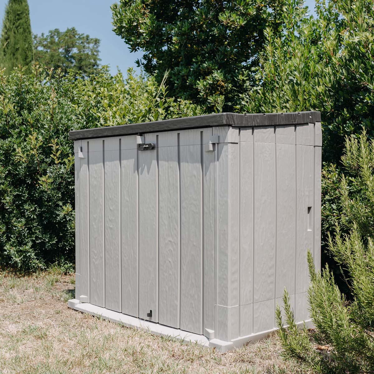 A grey plastic garden shed stands in front of green bushes on a lawn.