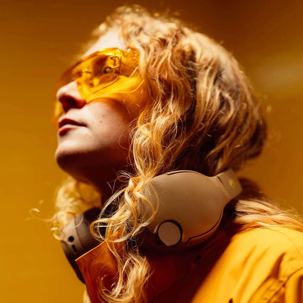 Persona con cabello rubio rizado, gafas de seguridad amarillas, auriculares alrededor del cuello y chaqueta amarilla.