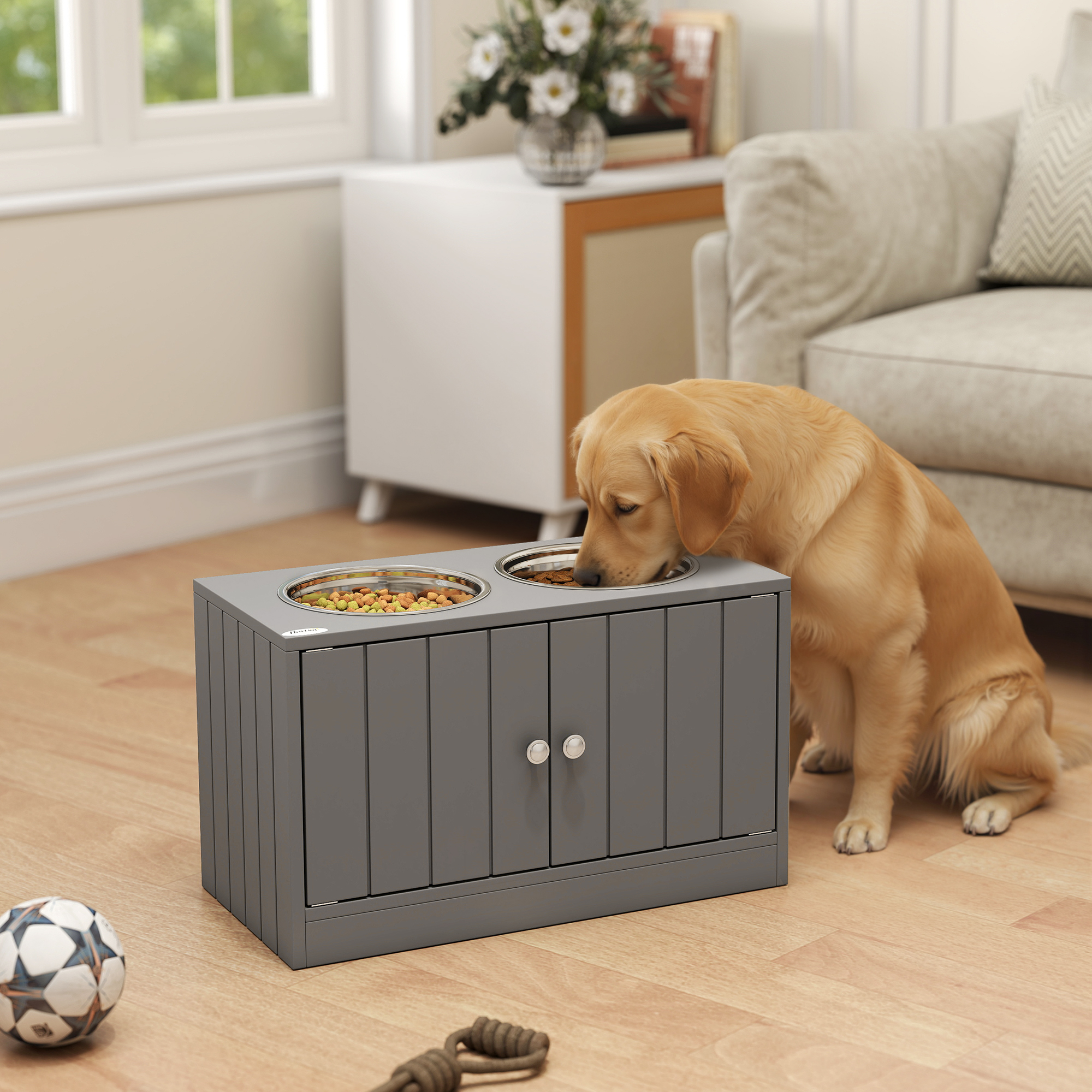 Un golden retriever come de dos cuencos en un armario gris en una sala con un balón de fútbol cerca.