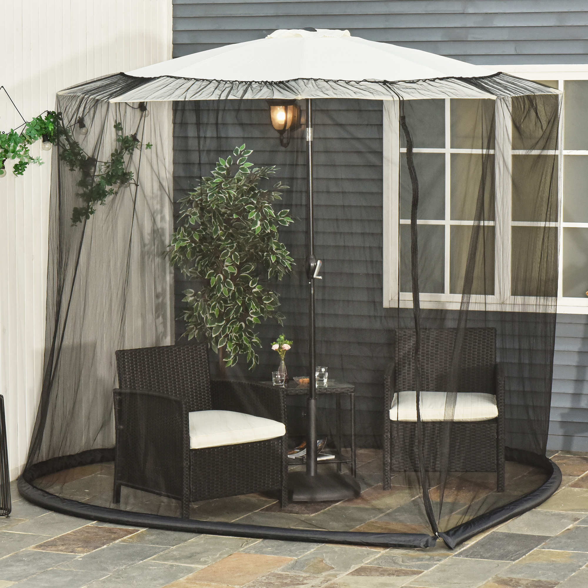 Una sombrilla blanca con mosquitera negra rodea dos sillones de ratán y una mesa pequeña en un patio de piedra.