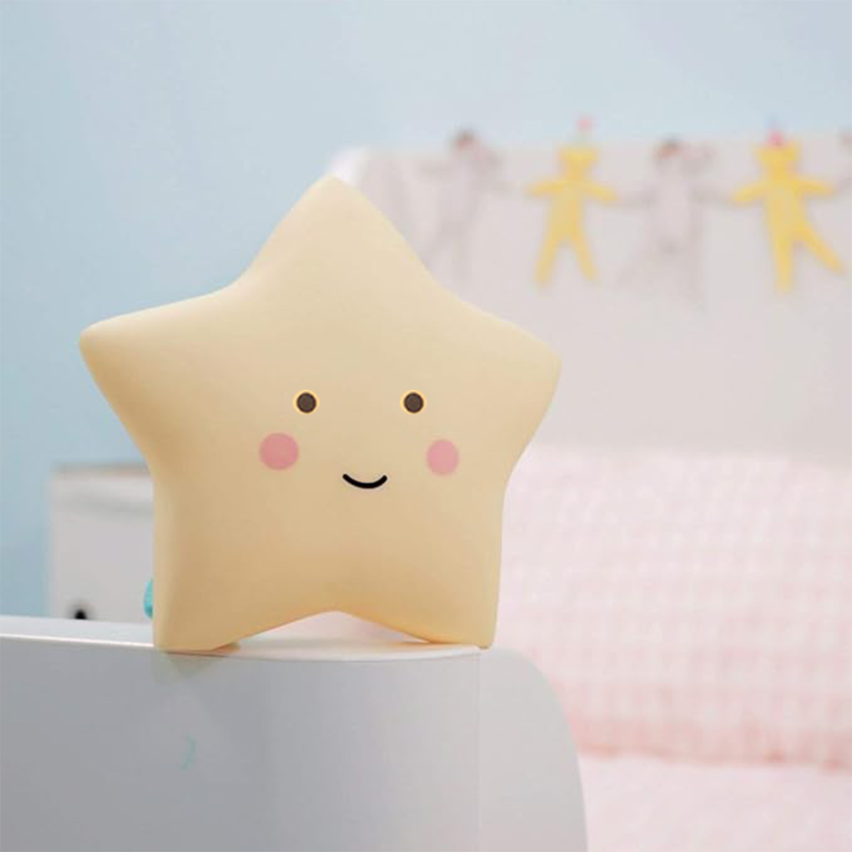 Eine gelbe, sternförmige Nachtlampe mit niedlichem Gesicht steht auf einem weißen Tisch vor einem verschwommenen Bett.