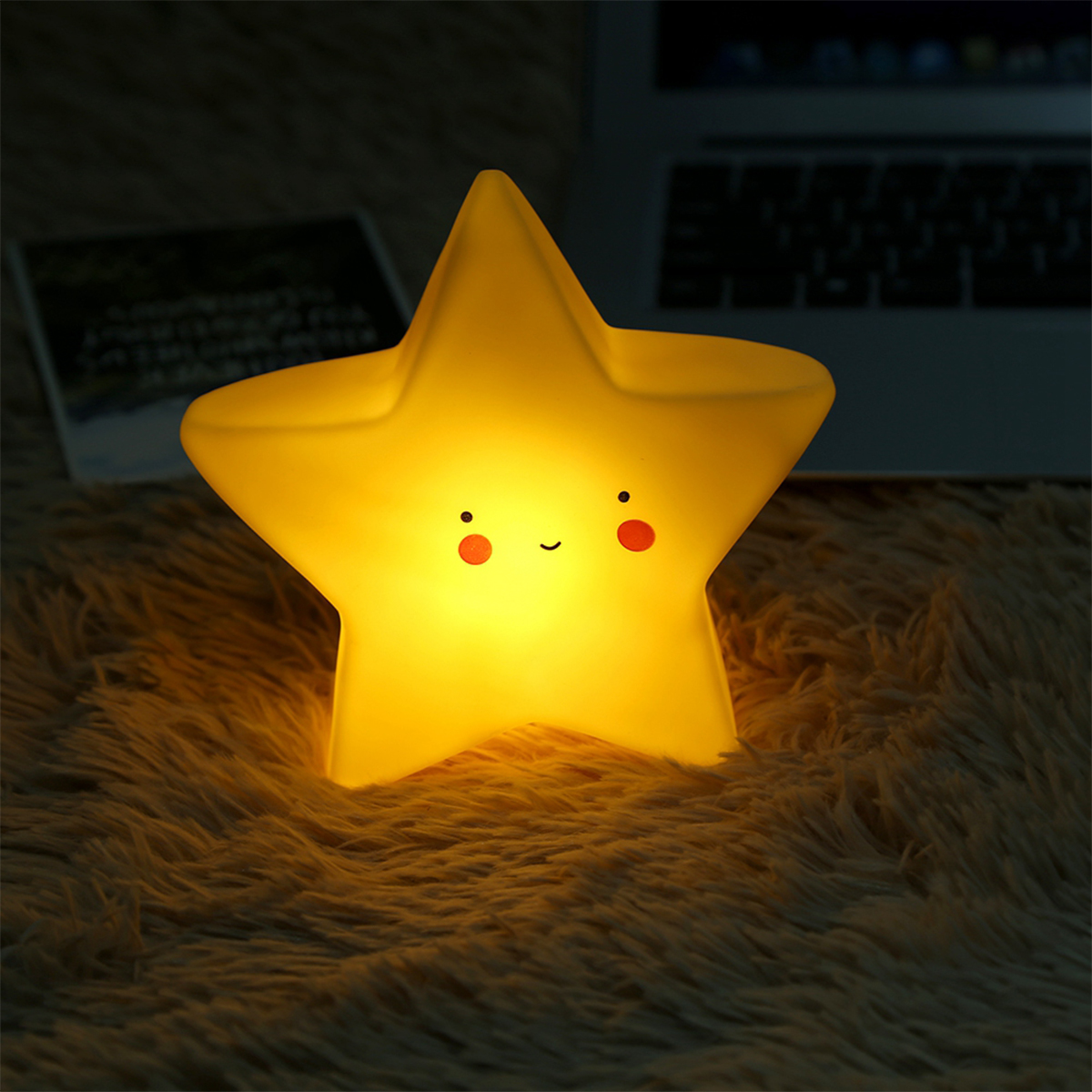 Eine leuchtende sternförmige Lampe mit süßem Gesicht sitzt auf einer flauschigen Decke neben einem Laptop.