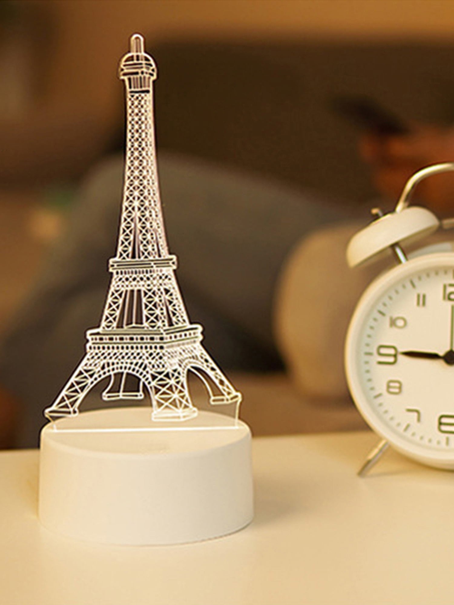 Eine weiß leuchtende Eiffel-Turm-Lampe auf einem Tisch neben einem weißen Wecker.