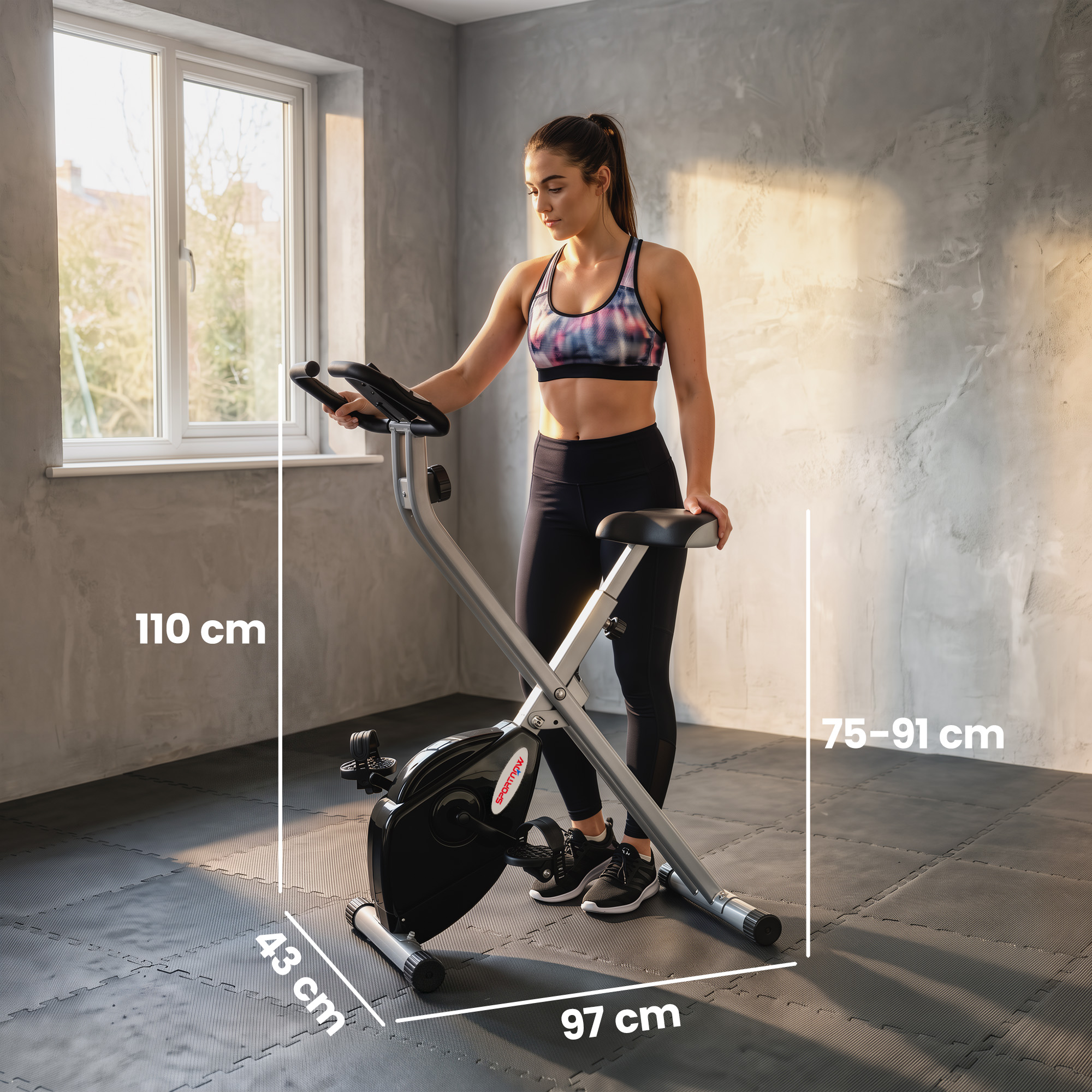 Una mujer con ropa deportiva está junto a una bicicleta estática plegable en una habitación con paredes grises.