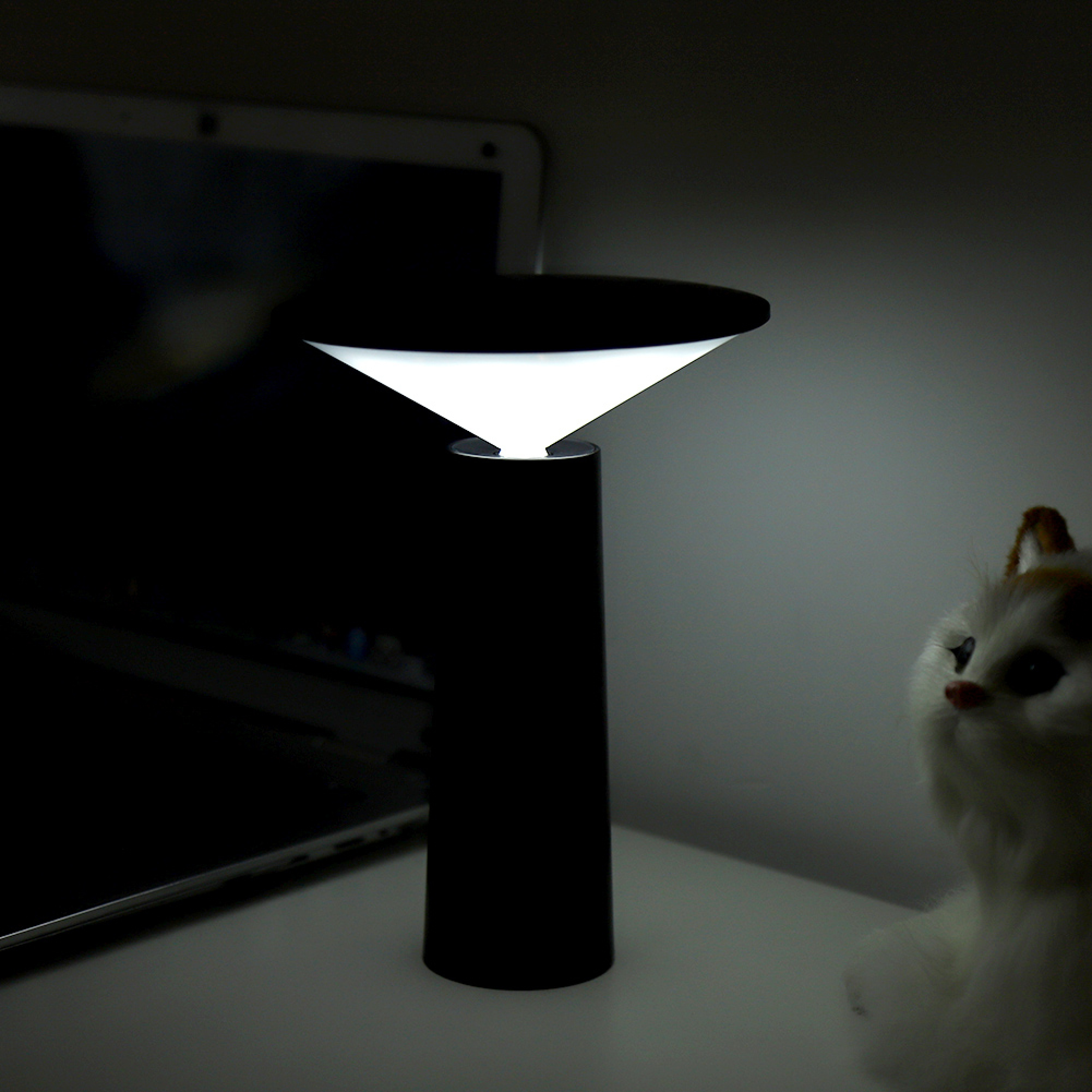 Eine moderne schwarze Tischlampe leuchtet auf einem Schreibtisch, neben einem Laptop und einer kleinen Stoffkatze.
