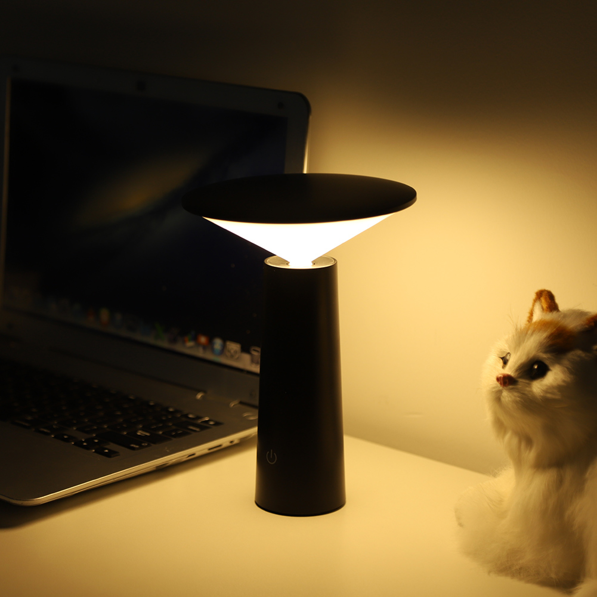 Eine schwarze, moderne Schreibtischlampe leuchtet sanft zwischen einem Laptop und einer kleinen weißen Katzendekoration.