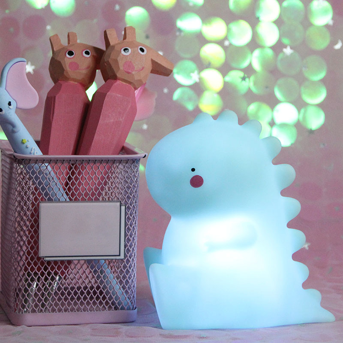 Ein Stiftehalter mit Holzfiguren und eine blaue Dinosaurier-Lampe vor einem pinken Hintergrund.