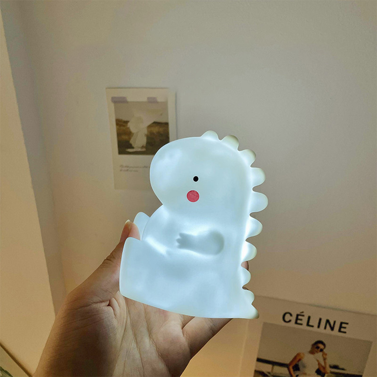 Eine Hand hält eine kleine, weiße Dinosaurier-Nachtlampe vor einer Wand.