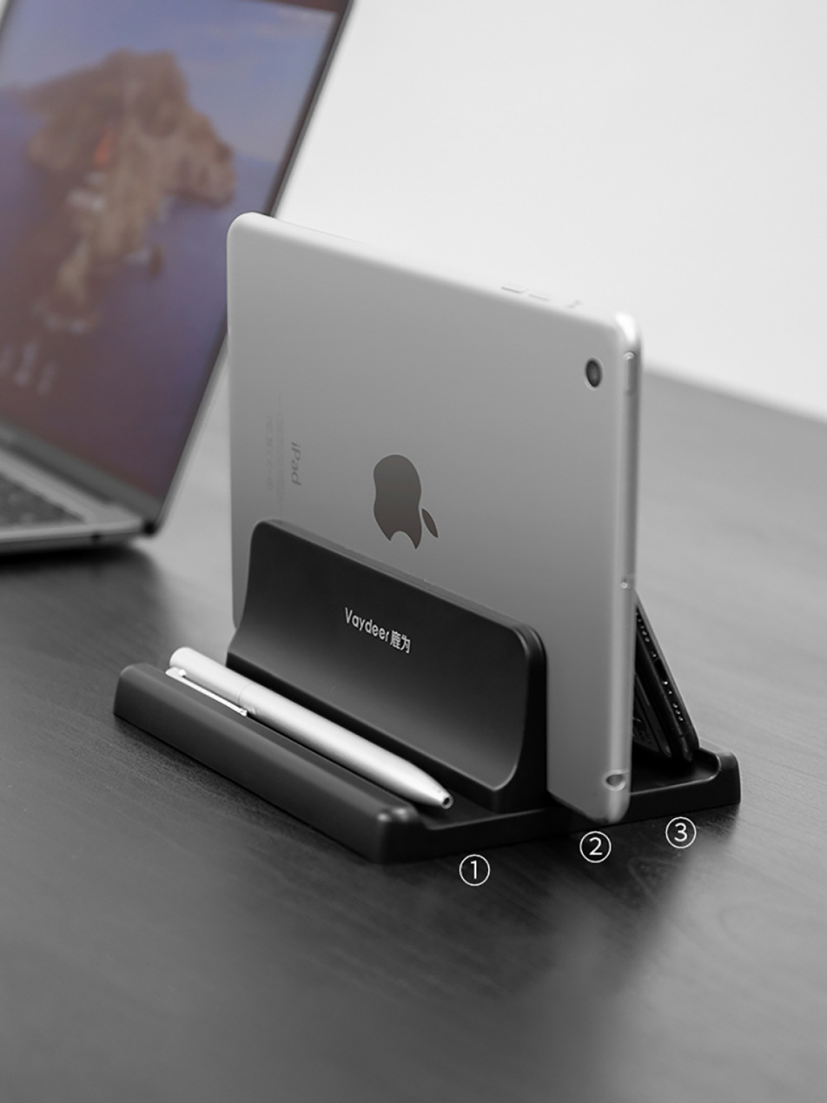 Ein schwarzer Ständer auf einem Tisch hält ein silbernes Tablet und einen Stift, mit einem Laptop im Hintergrund.