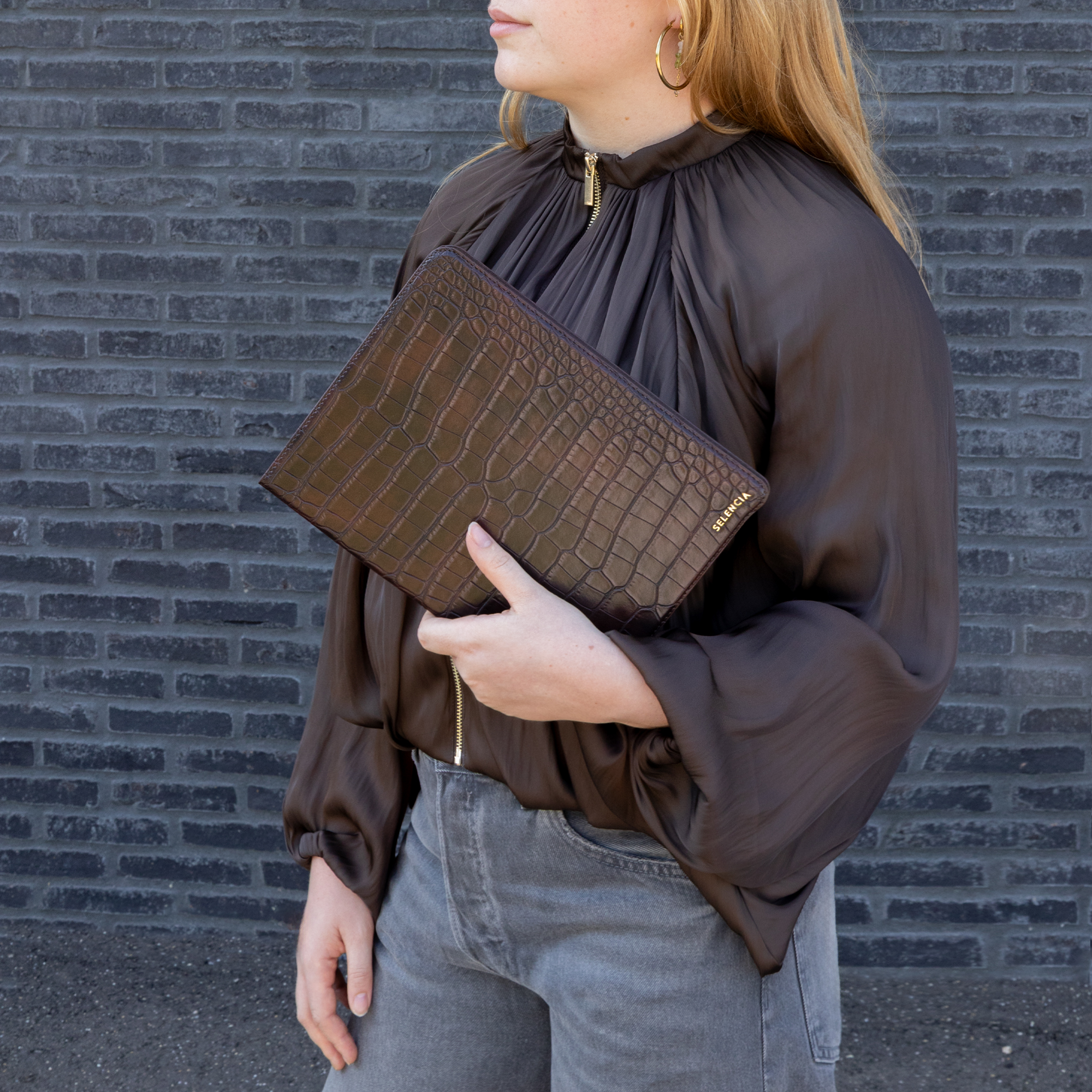 Een vrouw in een bruine blouse en grijze jeans houdt een bruine croco laptophoes voor een donkere bakstenen muur.