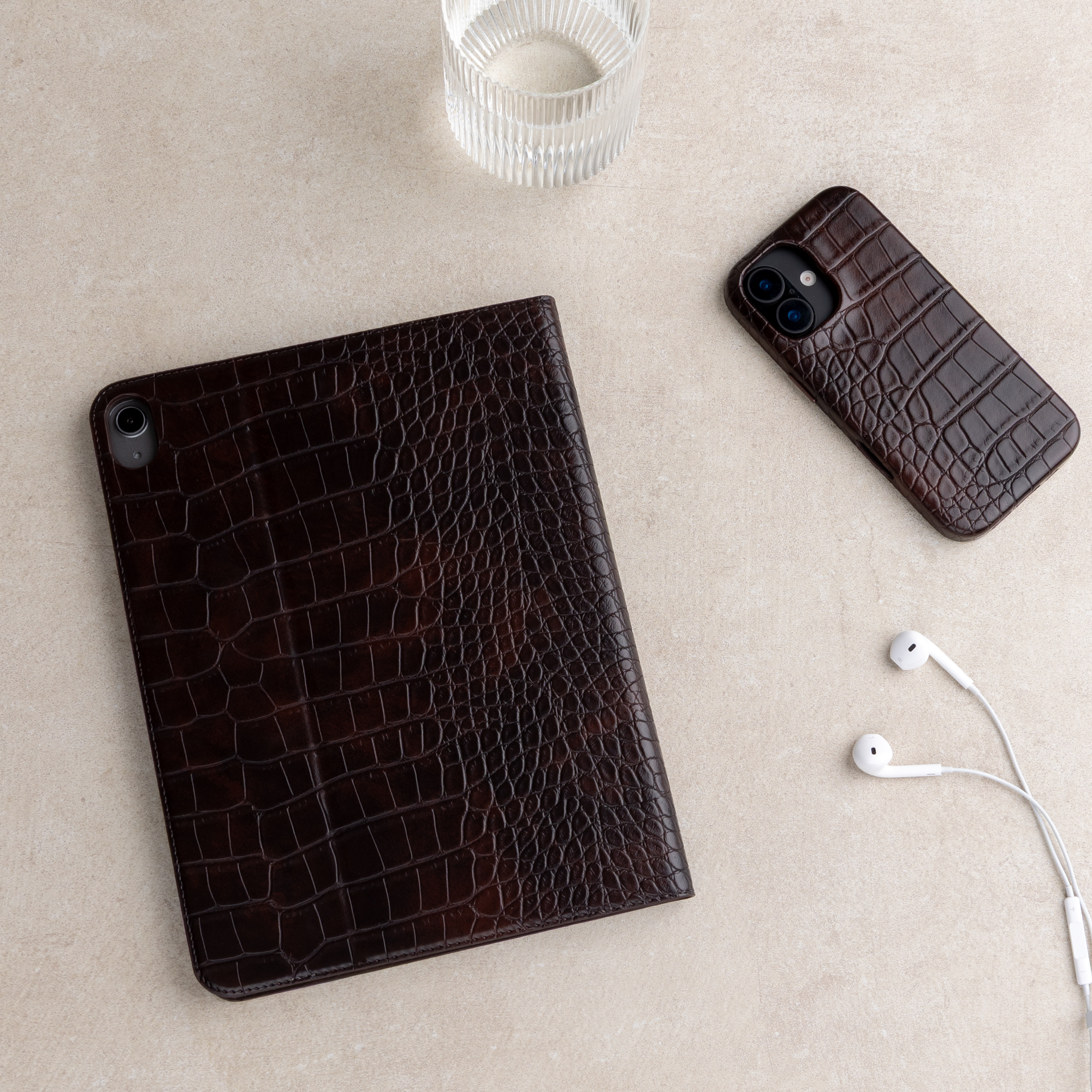 Bruine lederen hoezen voor tablet en smartphone, witte oortjes en een glas op beige ondergrond.
