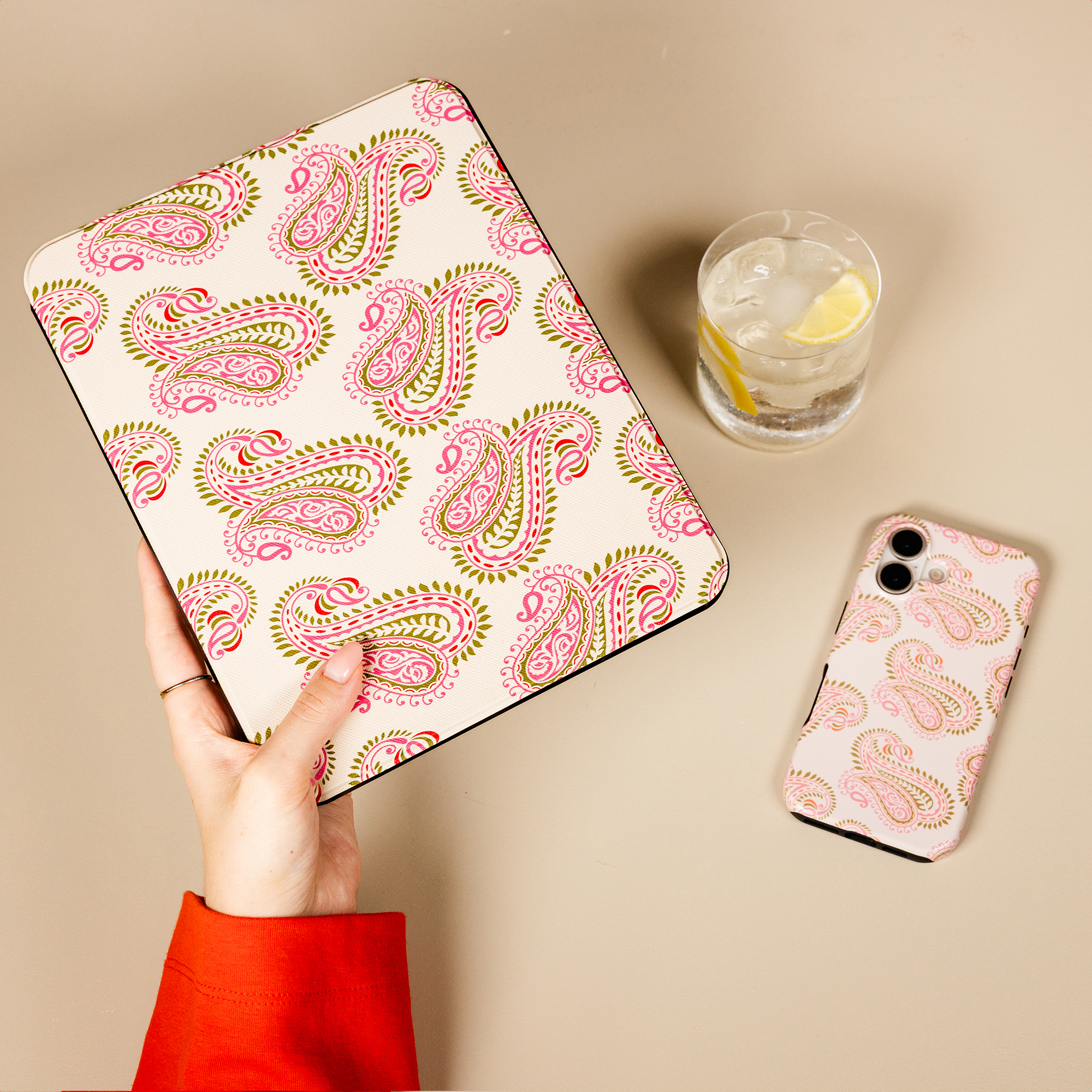 Une main tient une housse rose à motif cachemire avec une coque assortie et un verre d'eau citronnée sur fond beige.
