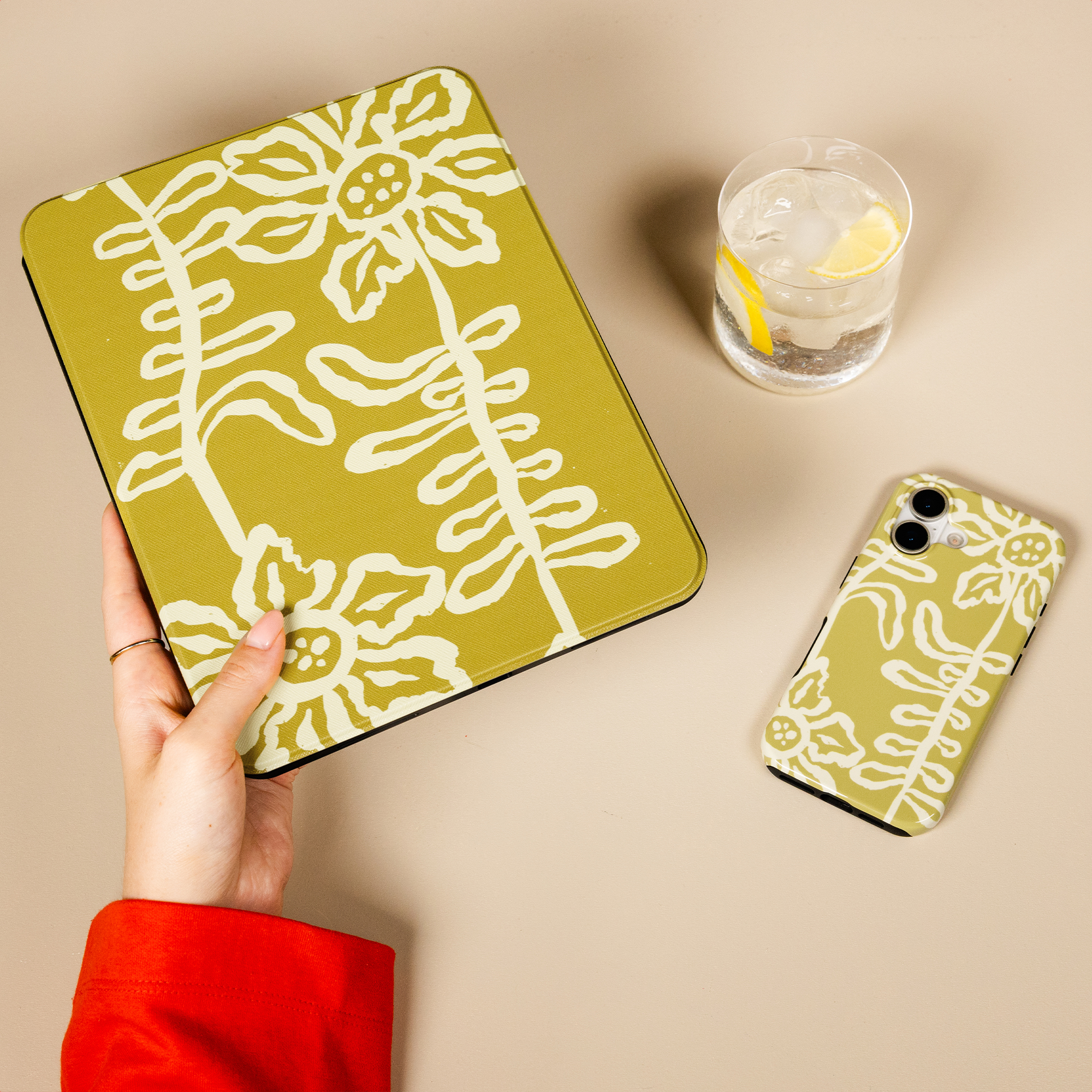 Une main en pull rouge tient une housse de tablette vert olive à fleurs, près d'un étui de téléphone assorti et d'un verre.