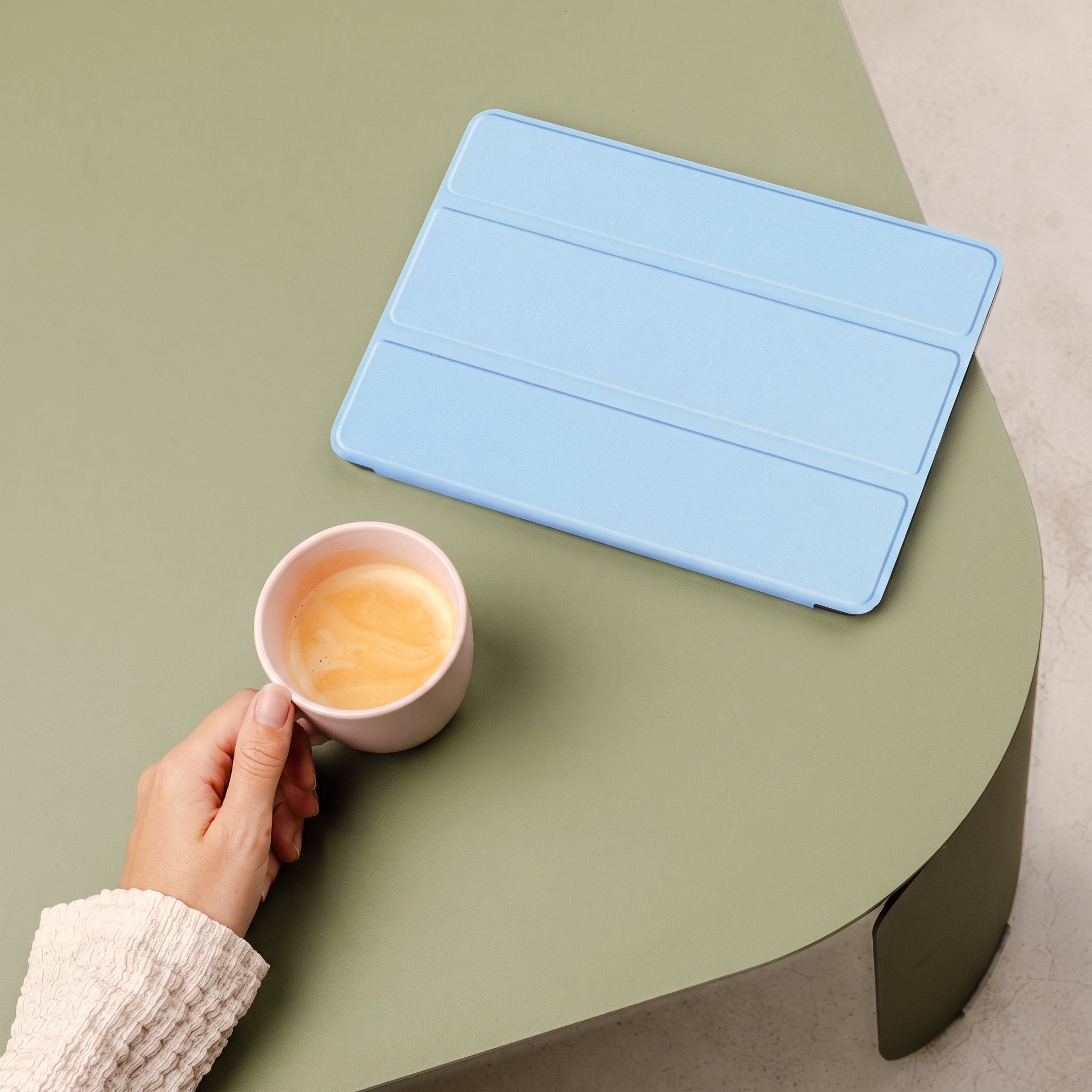Een hand houdt een roze kopje koffie vast naast een blauwe tablethoes op een groene tafel.