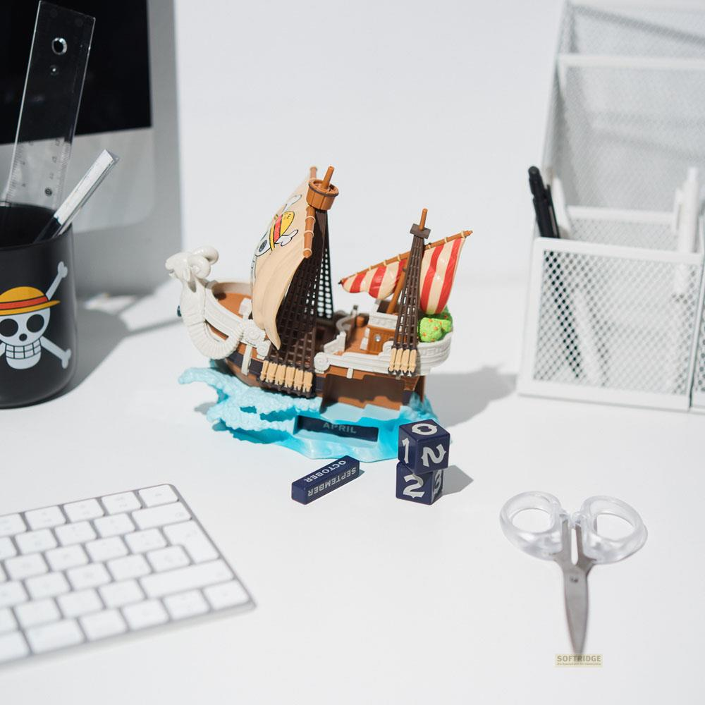 Un juguete de barco pirata en miniatura se encuentra en un escritorio blanco con teclado, tijeras y bloque de calendario.
