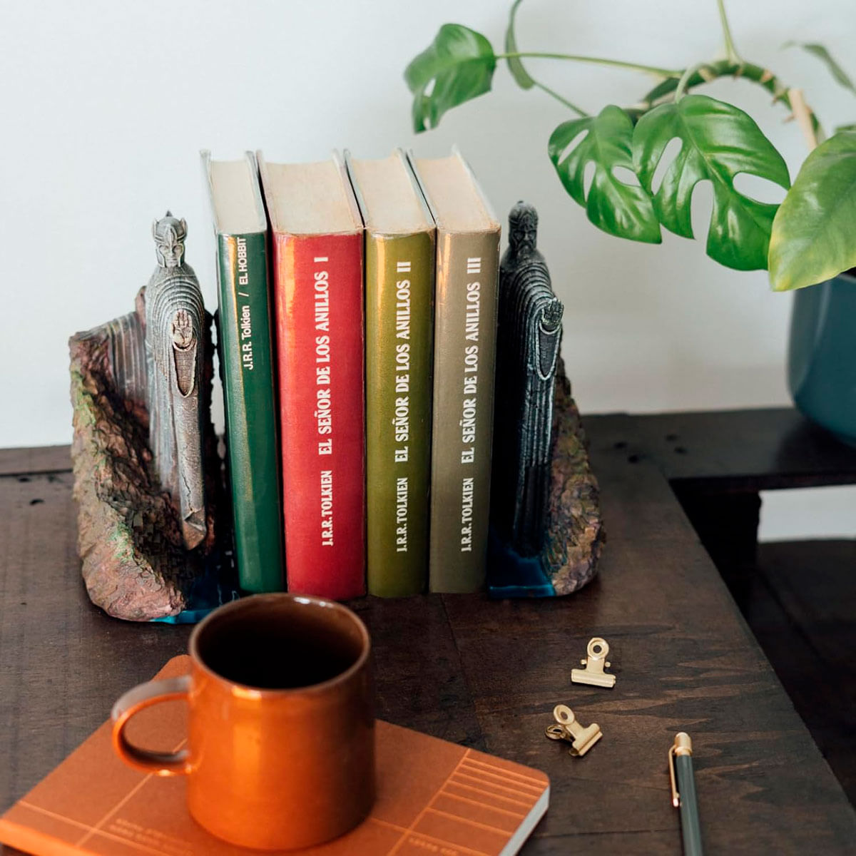 Libros de El Señor de los Anillos entre sujetalibros de estatuas en escritorio con taza naranja.