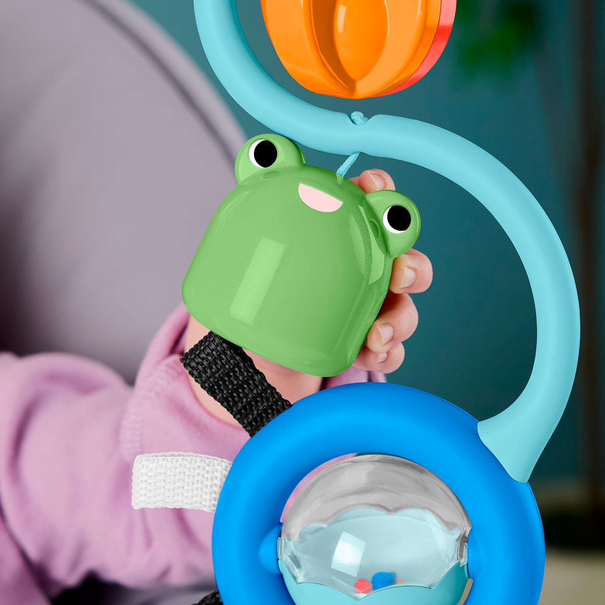 La mano de un bebé sostiene un juguete verde con forma de rana que cuelga de un arco azul.