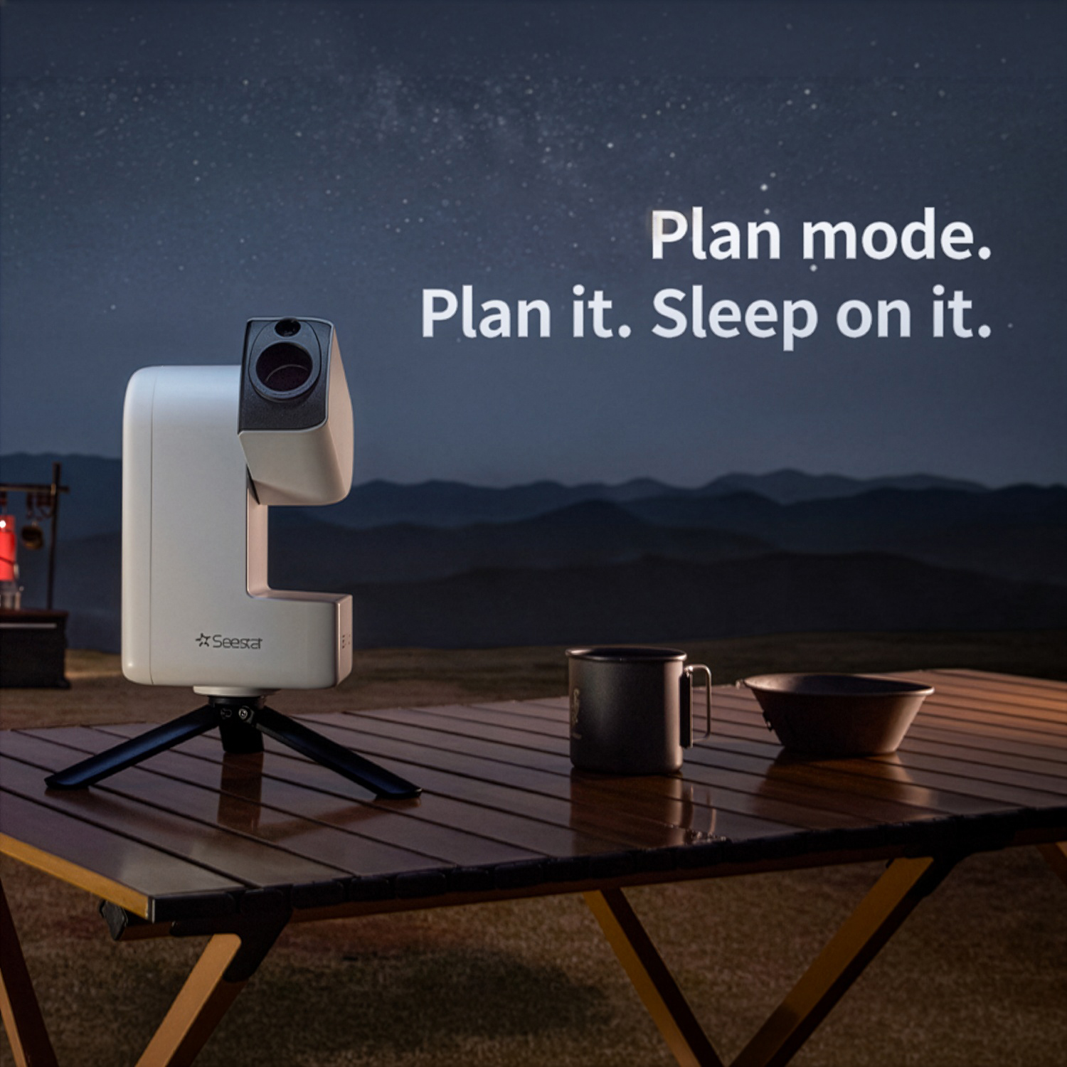 Un telescopio Seestar y una taza en una mesa de madera al aire libre de noche con montañas y cielo estrellado.