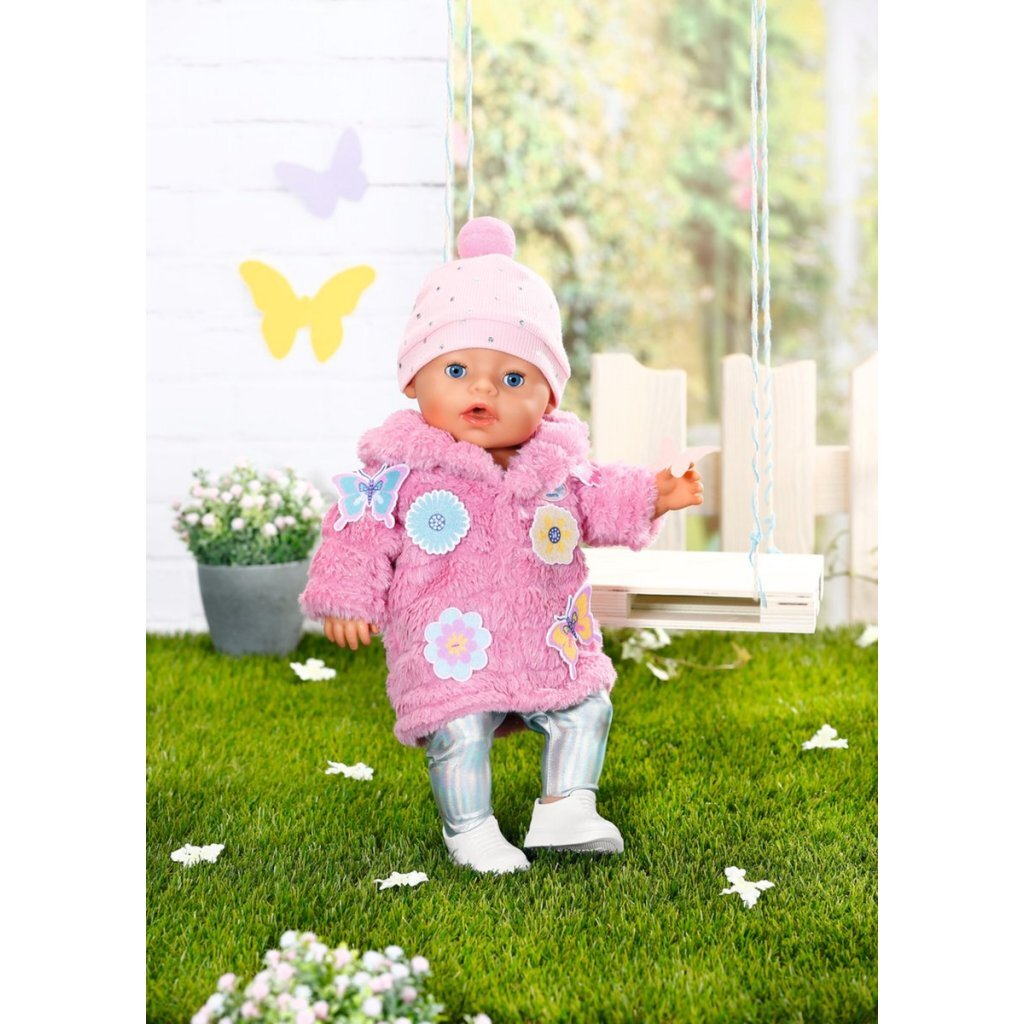 Un muñeco bebé con abrigo rosa esponjoso y gorro está de pie sobre césped verde ante un columpio y una pared blanca.