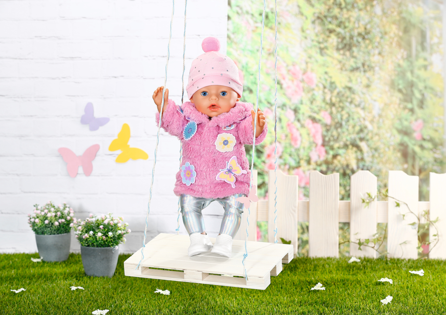 Una muñeca con abrigo peludo rosa y gorro está de pie en un columpio blanco frente a un jardín.