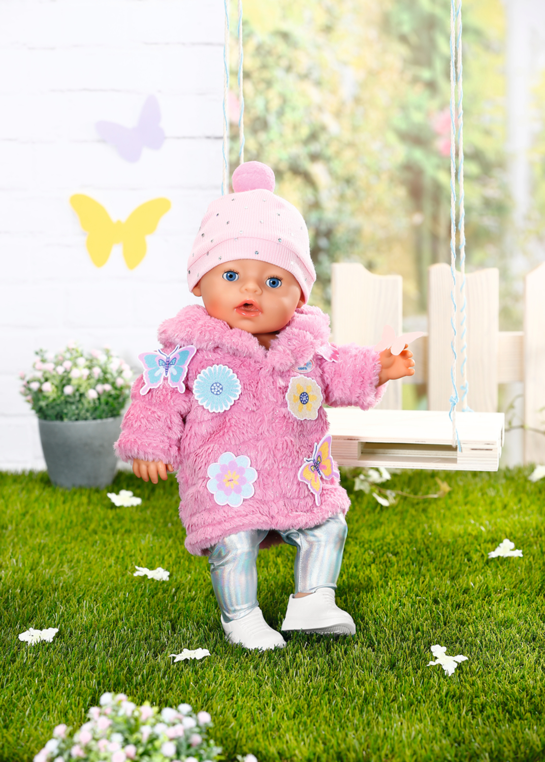 Un muñeco bebé con abrigo rosa, gorro y mallas brillantes está de pie en el césped frente a un columpio.