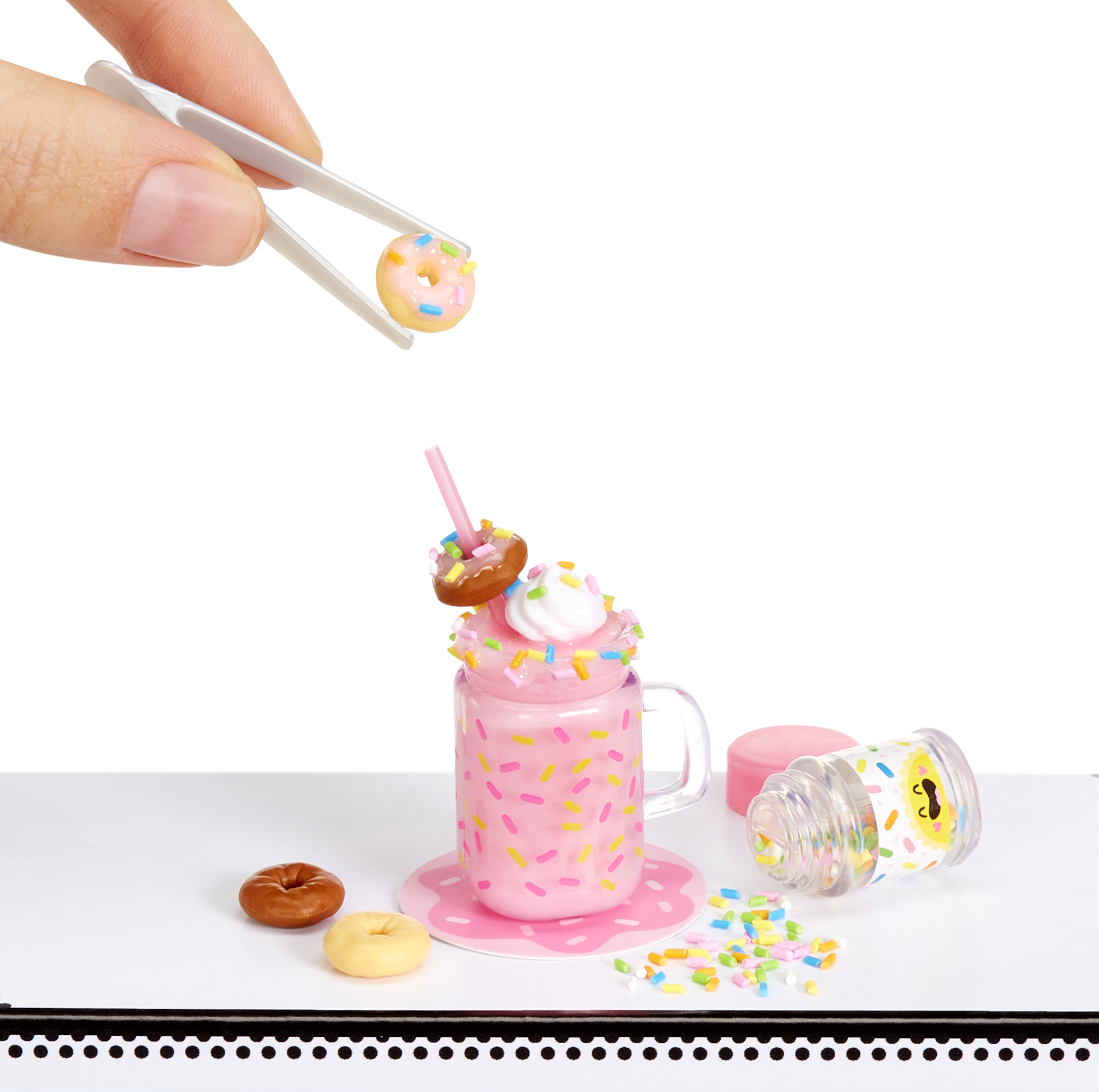 Unas pinzas colocan un minidona en un batido rosa decorado con chispas, crema batida y otra dona.