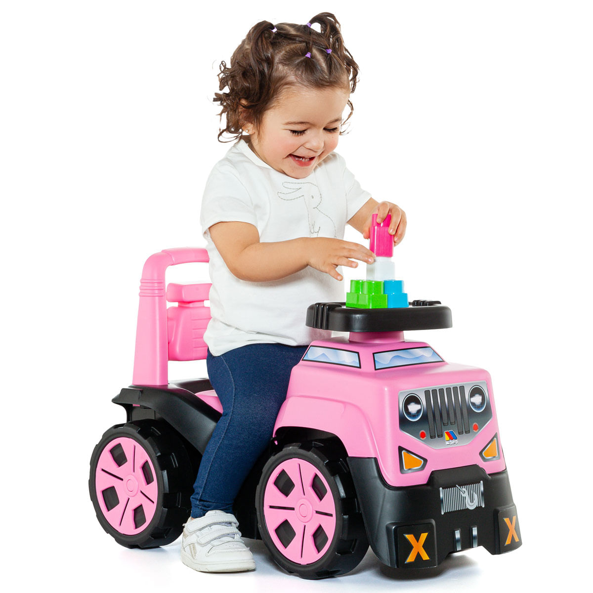 Una niña pequeña sonriente está sentada en un coche de juguete rosa, apilando bloques de colores en el volante.