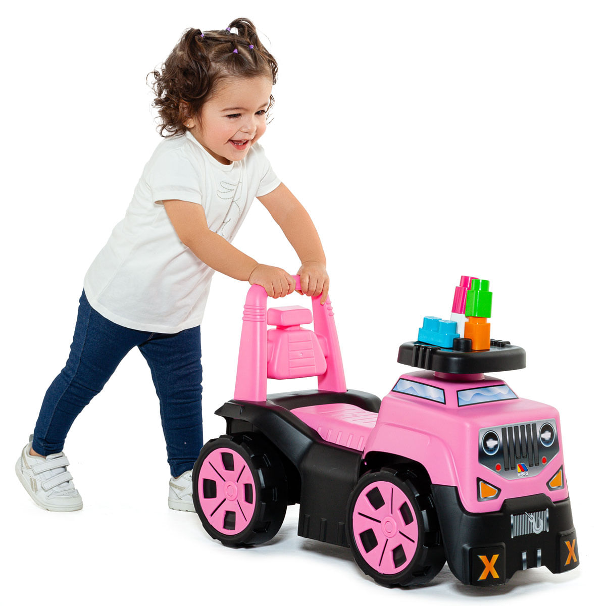 Un niño pequeño sonriente empuja un camión de juguete rosa y negro cargado con bloques coloridos sobre fondo blanco.