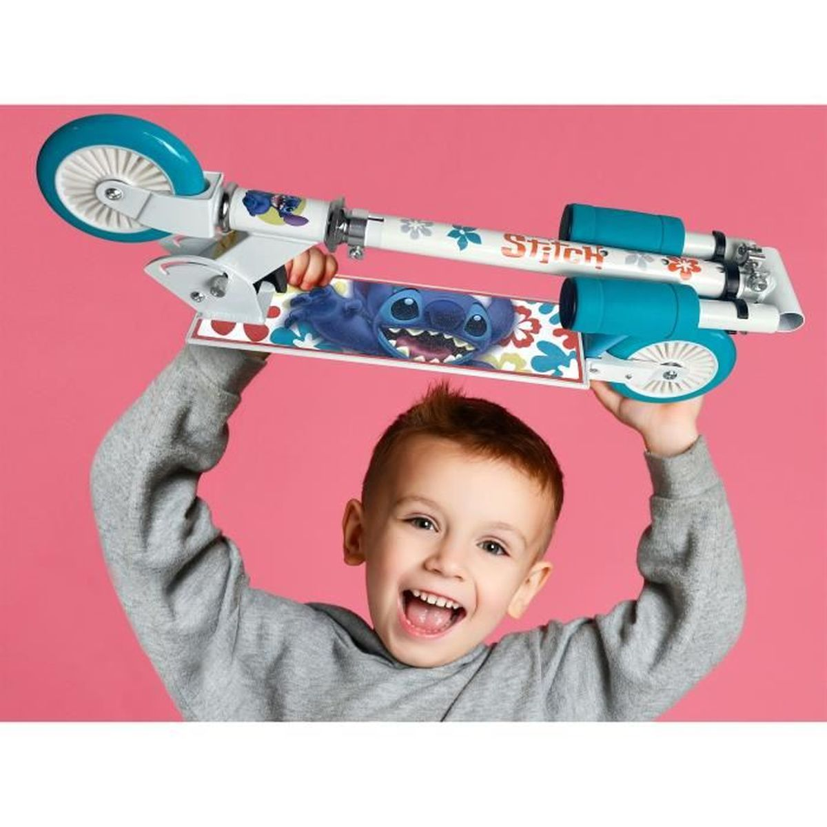 Un niño feliz con jersey gris sostiene un patinete de Stitch blanco y azul sobre su cabeza ante un fondo rosa.